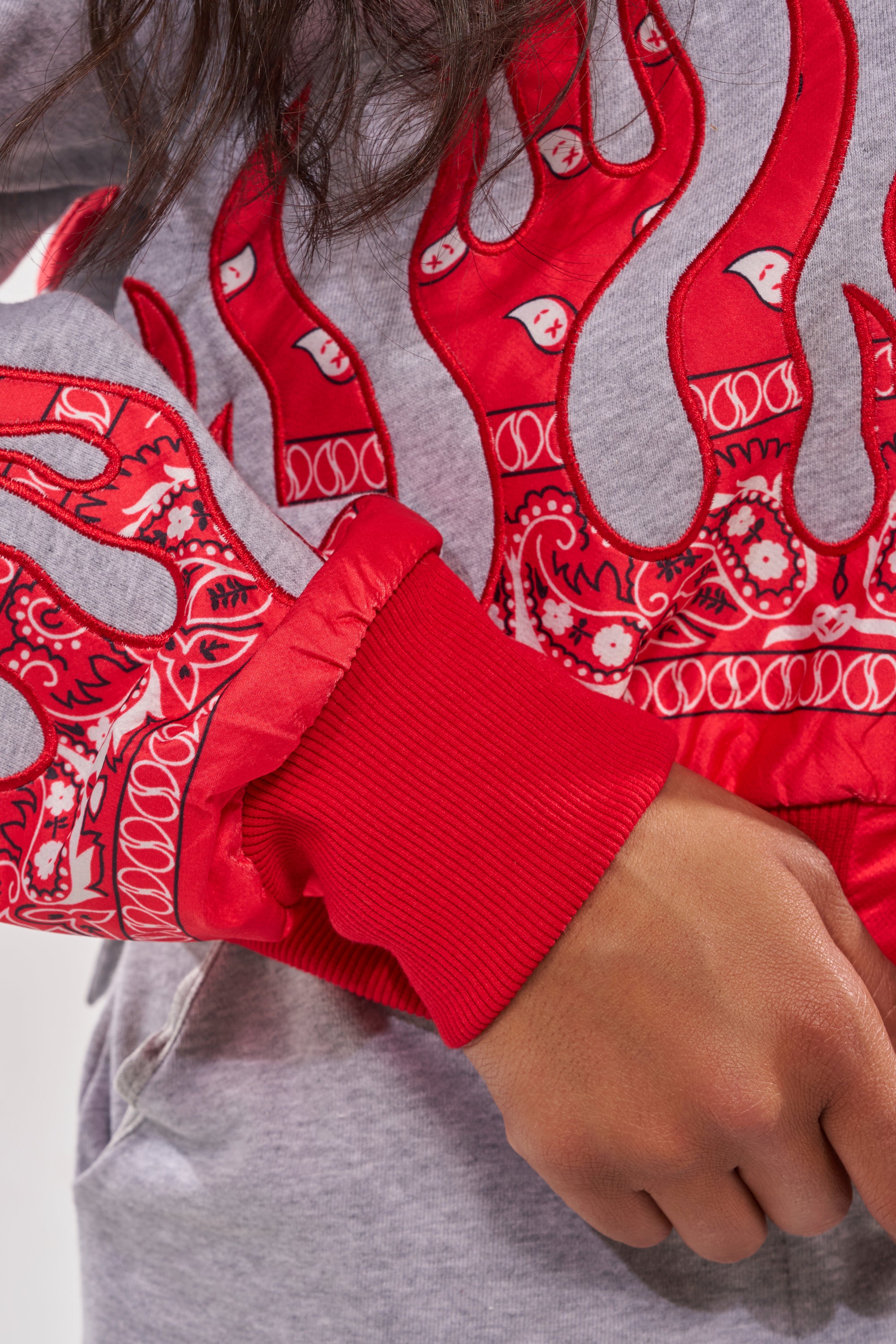A person wearing the FIRE TO THE FLAMES SWEATSHIRT—a gray street-style hoodie with bold red paisley, embroidered flame graphics, and bright red ribbed cuffs—rests their hand near the pocket. 10