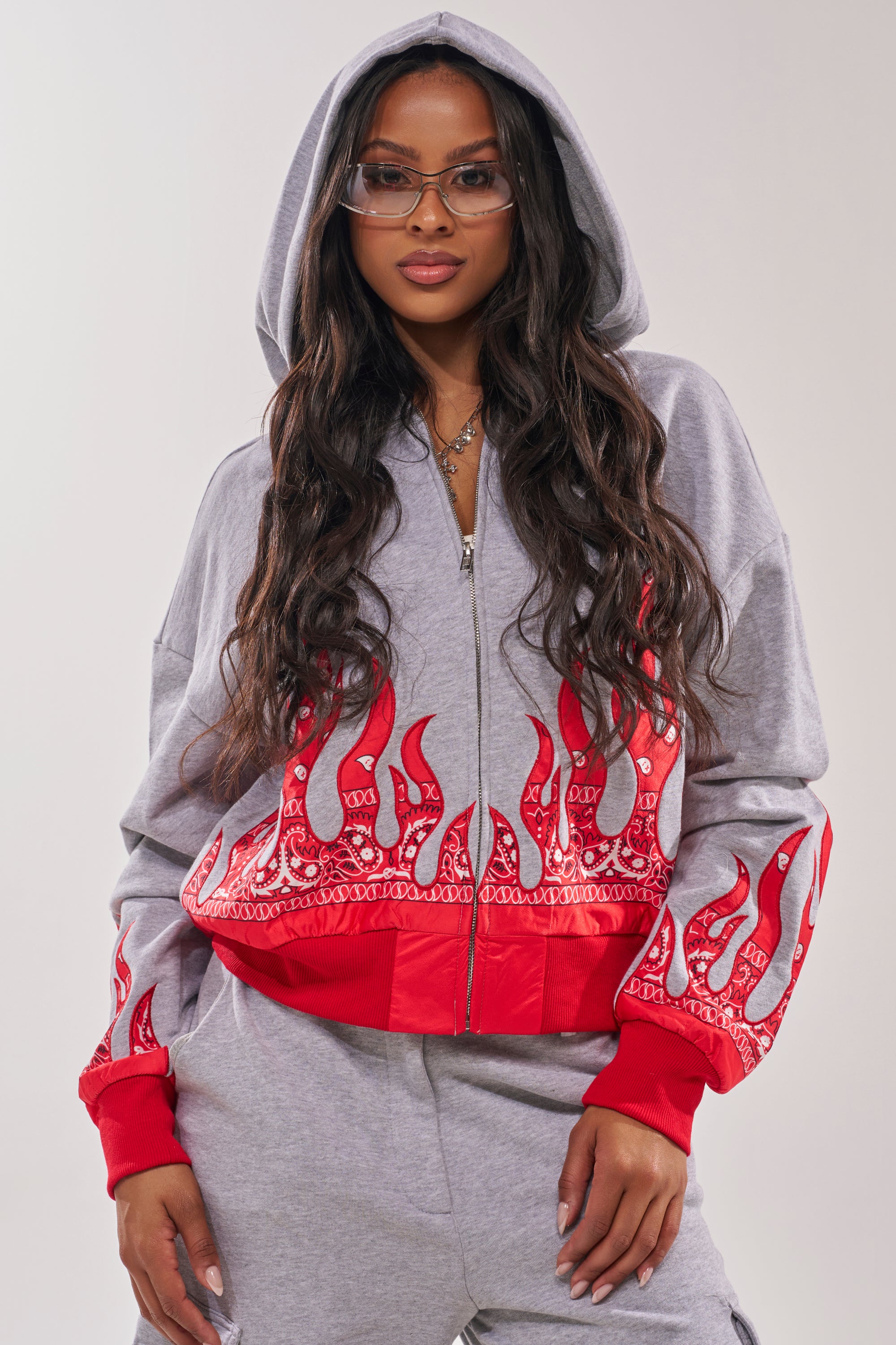 A woman poses confidently in the FIRE TO THE FLAMES SWEATSHIRT, a gray street-style hoodie with red embroidered flame designs on the sleeves and waistband. She completes the bold look with long wavy hair and glasses. 3