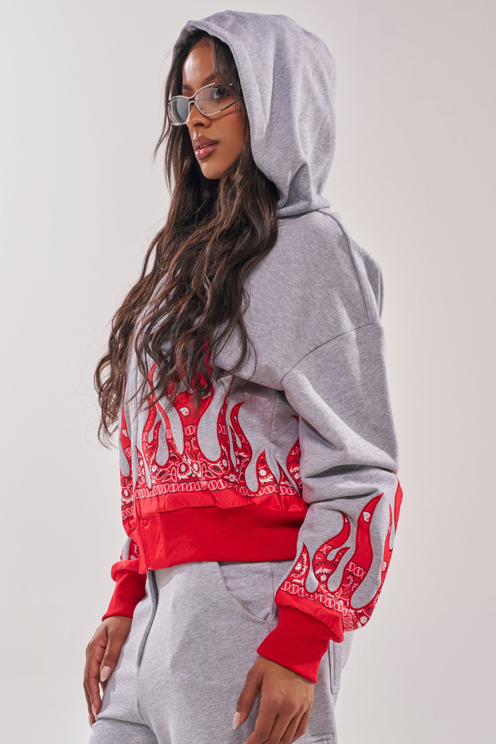 A woman with long, wavy hair and clear glasses poses in profile against a plain background, wearing the FIRE TO THE FLAMES SWEATSHIRT—a gray street-style hoodie with embroidered flame graphics. 4