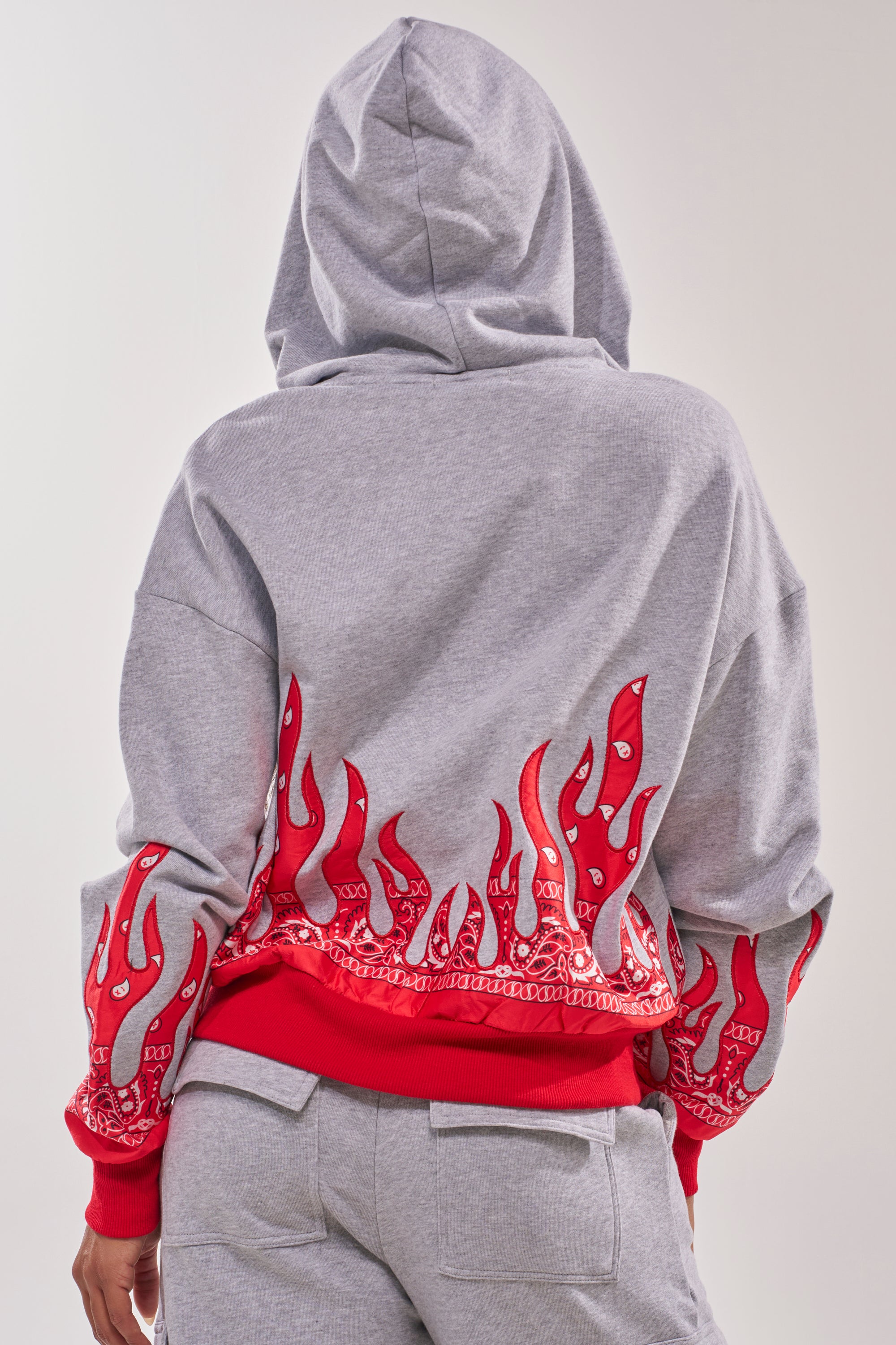 Someone faces away from the camera wearing the FIRE TO THE FLAMES SWEATSHIRT, a gray street-style hoodie featuring a red flame and bandana pattern on the back and sleeves, paired with matching gray sweatpants. 5
