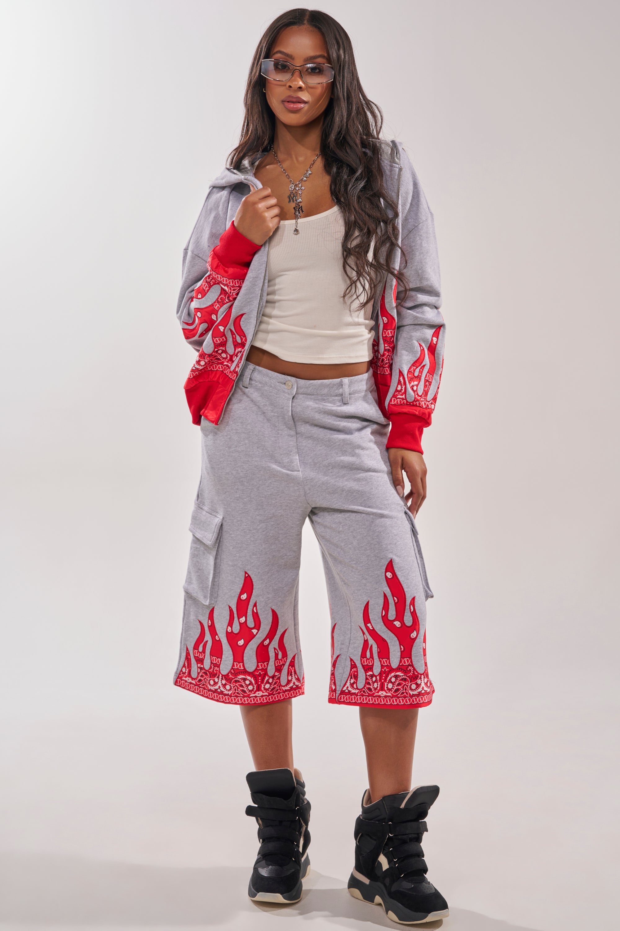 A woman in the FIRE TO THE FLAMES SWEATSHIRT, matching shorts, white tank top, black high-top sneakers, and sunglasses stands against a plain background. 6