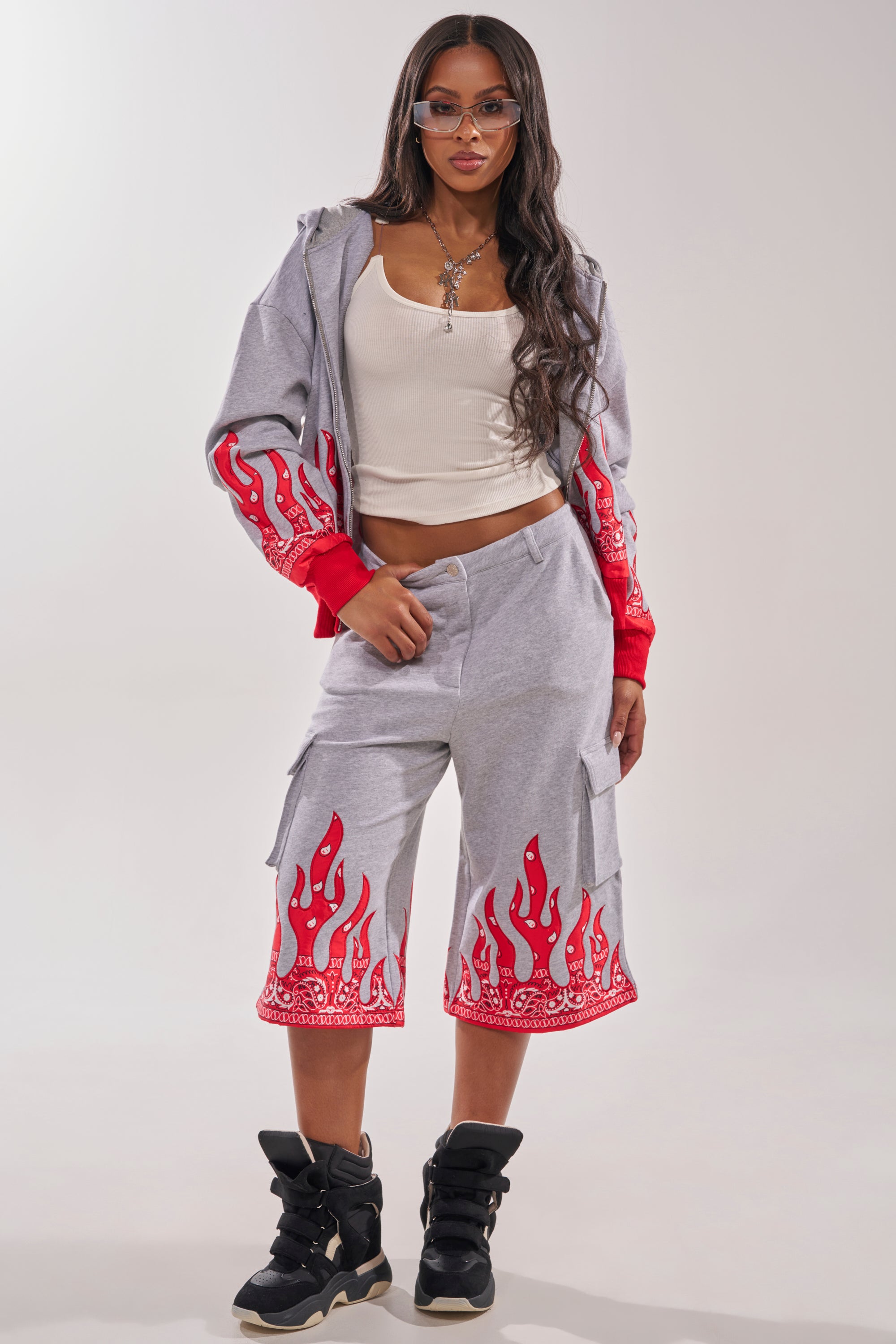 A woman with long hair wears the FIRE TO THE FLAMES SWEATSHIRT and matching shorts with flame embroidery, a white tank top, sunglasses, a necklace, and black high-top sneakers while standing against a plain background. 8