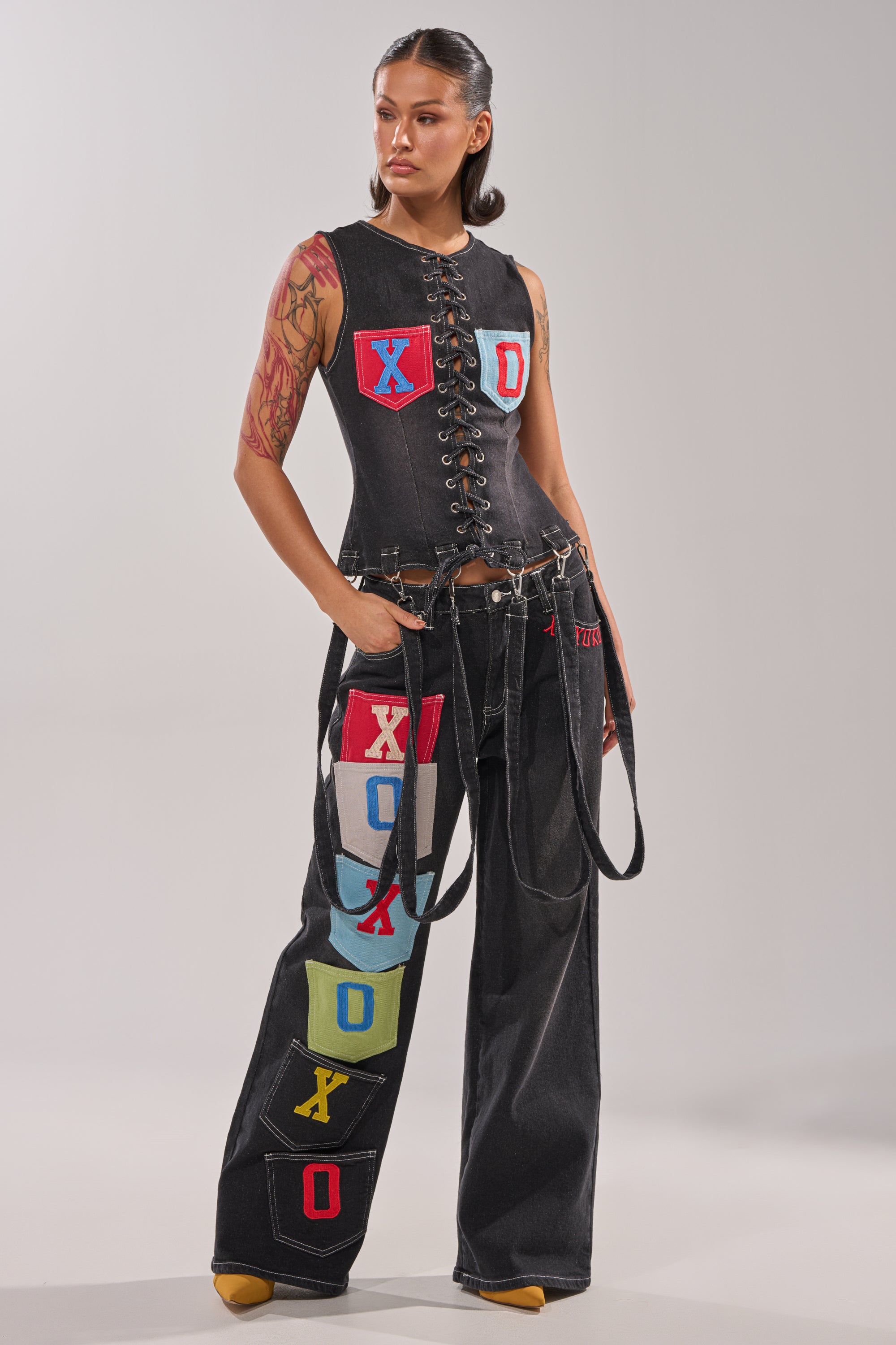 A woman stands confidently with hands in her pockets, wearing a sleeveless, lace-up black denim top and BRING IT BACK DENIM PATCHWORK PANT—black wide-leg jeans with colorful XOXO patch pockets—against a plain background. 0