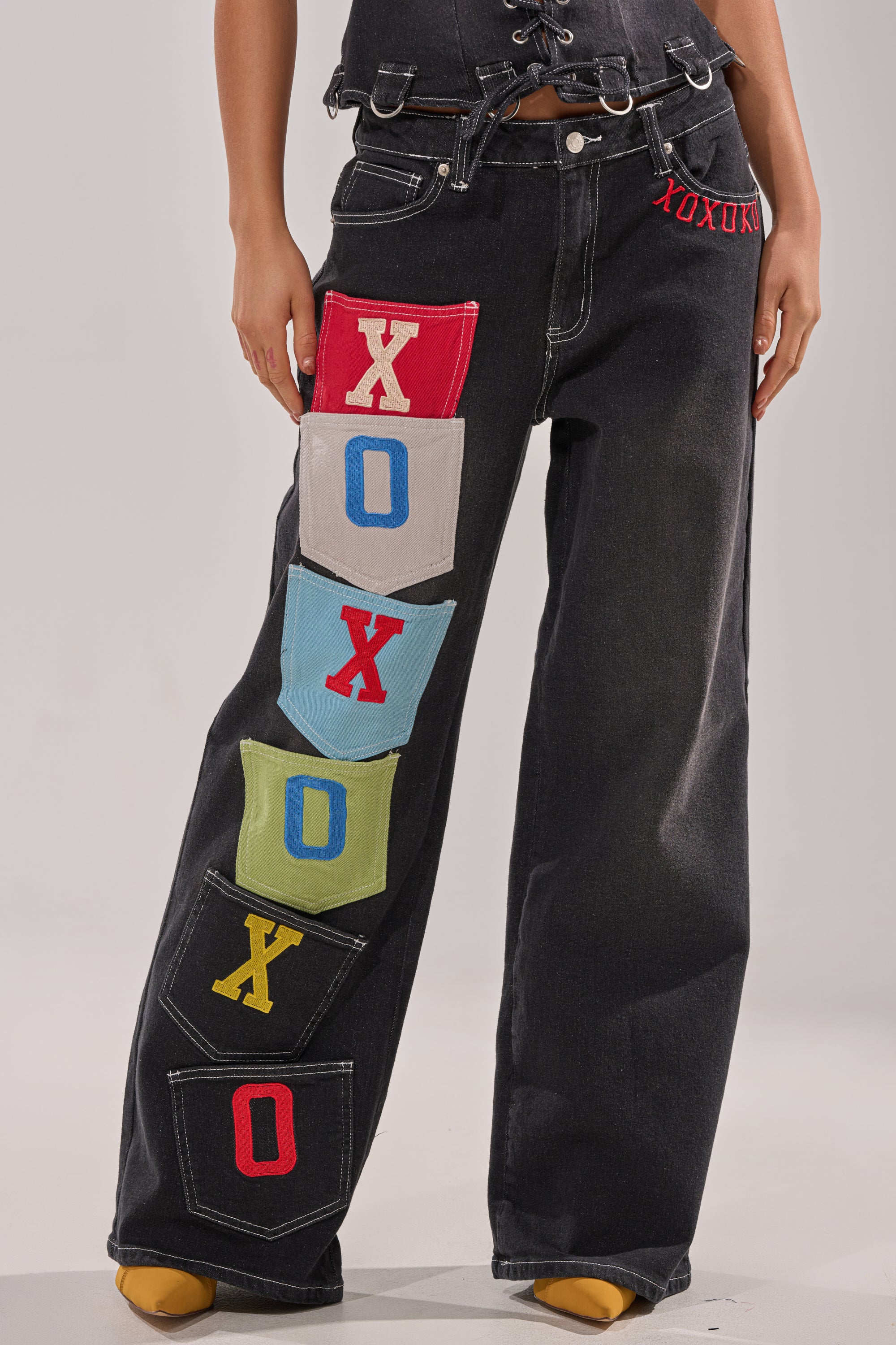 Wearing the BRING IT BACK DENIM PATCHWORK PANT—black wide-leg jeans with colorful cargo pockets on one leg spelling "XOXOXO"—styled with mustard yellow shoes. 1