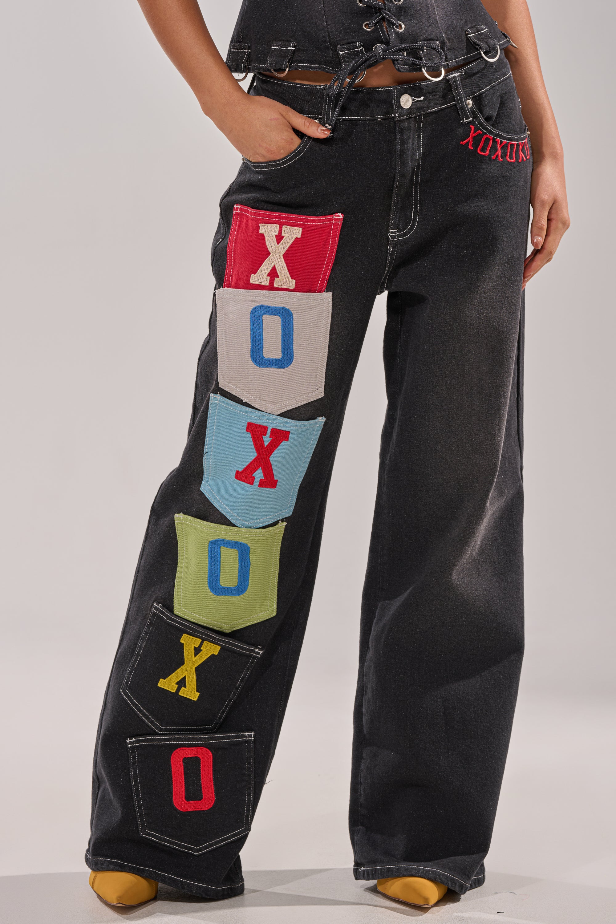 A person wears the BRING IT BACK DENIM PATCHWORK PANT—black wide-leg jeans with colorful cargo patchwork pockets spelling "XOXOX" vertically on one leg—paired with a black top and yellow shoes. 2