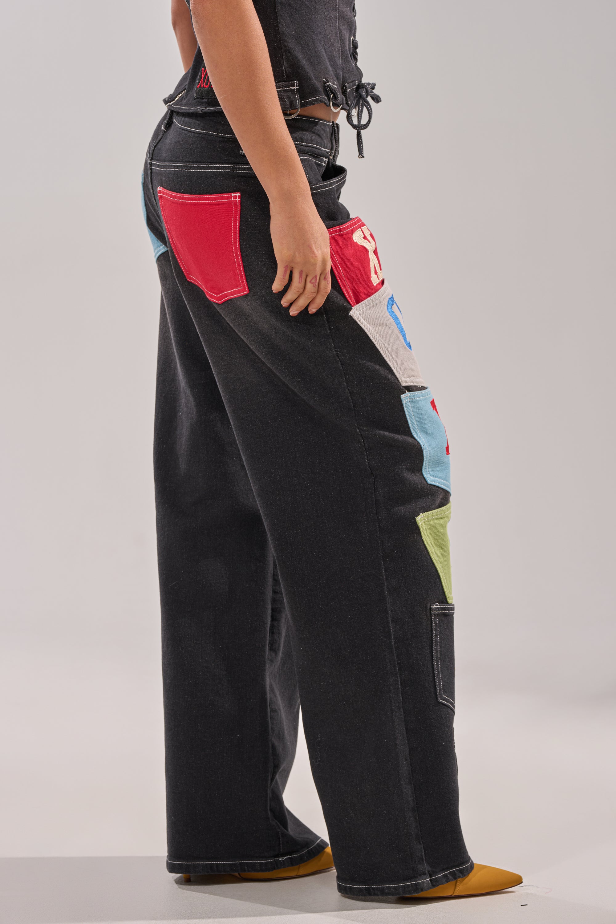 A person models the BRING IT BACK DENIM PATCHWORK PANT—black wide-leg street-style jeans with colorful cargo patchwork pockets in red, blue, green, white, and gray—paired with orange pointed-toe shoes against a plain light backdrop. 3