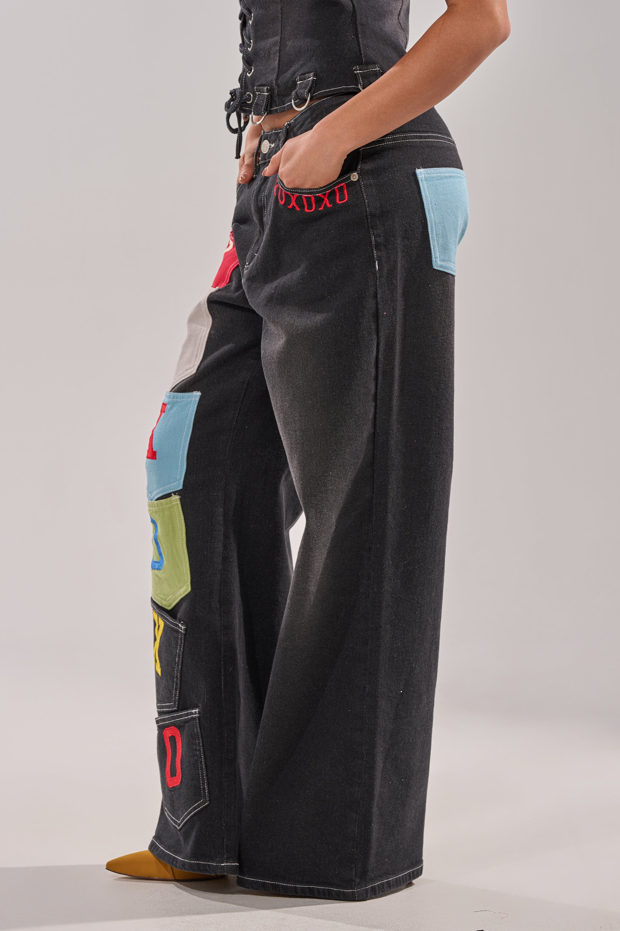 A person stands with one hand in their pocket, wearing the BRING IT BACK DENIM PATCHWORK PANT—black wide-leg street-style jeans with colorful patchwork and large blue pockets—paired with a black corset-style top. 4