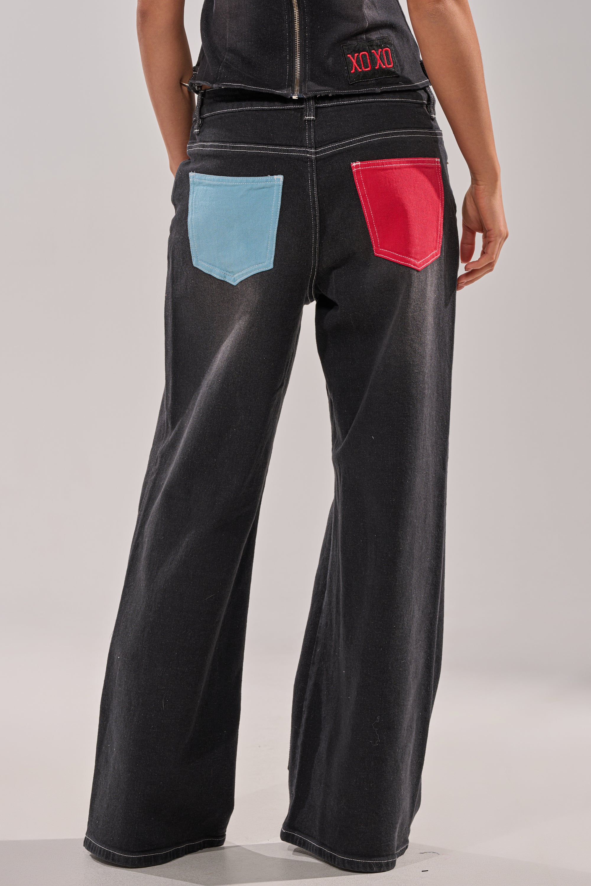 Model wears the BRING IT BACK DENIM PATCHWORK PANT—black wide-leg jeans with a light blue left pocket, red right pocket, and bold red "XO XO" on the upper back—making a standout street-style statement against a neutral background. 5