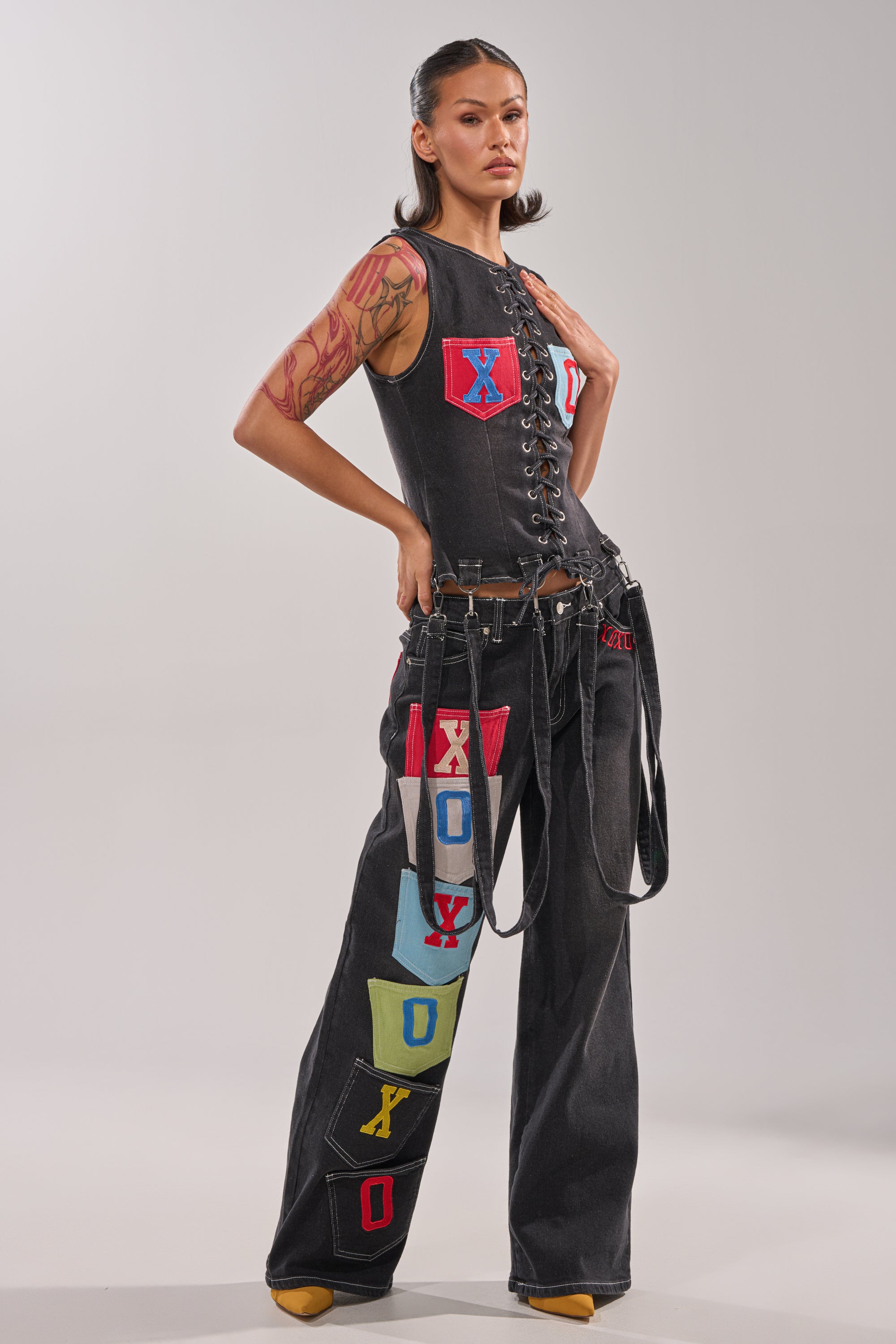 A woman poses confidently in the BRING IT BACK DENIM PATCHWORK PANT, black wide-leg jeans adorned with colorful "XOXO" letter patches. She pairs them with a black lace-up sleeveless top, has slicked-back hair, tattoos, and yellow shoes. 6