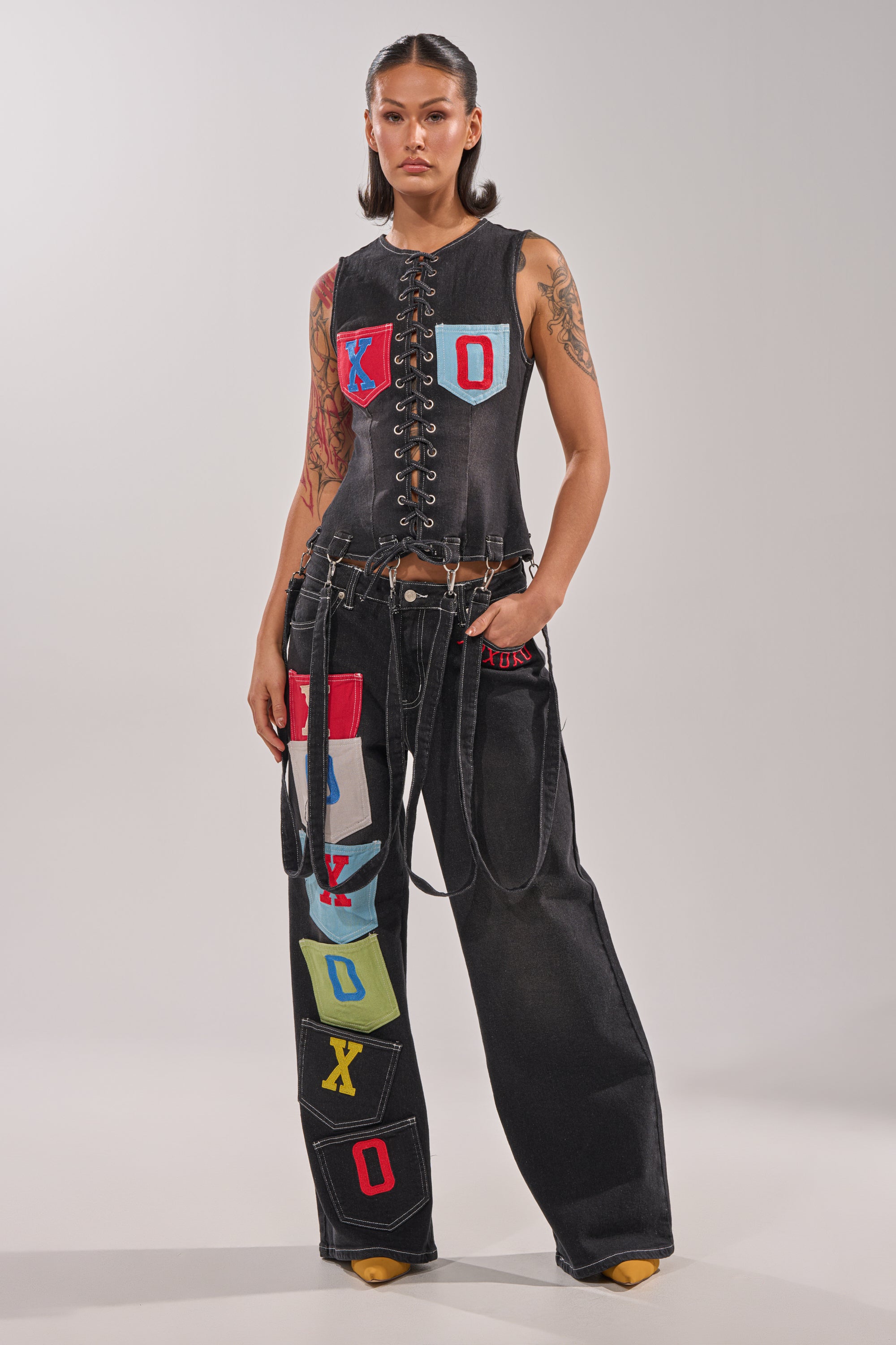 A woman stands confidently in the BRING IT BACK DENIM PATCHWORK PANT—black wide-leg street-style jeans featuring colorful patches and decorative chains—paired with a black lace-up vest with "XOXO" letters and yellow pointed-toe shoes. 7