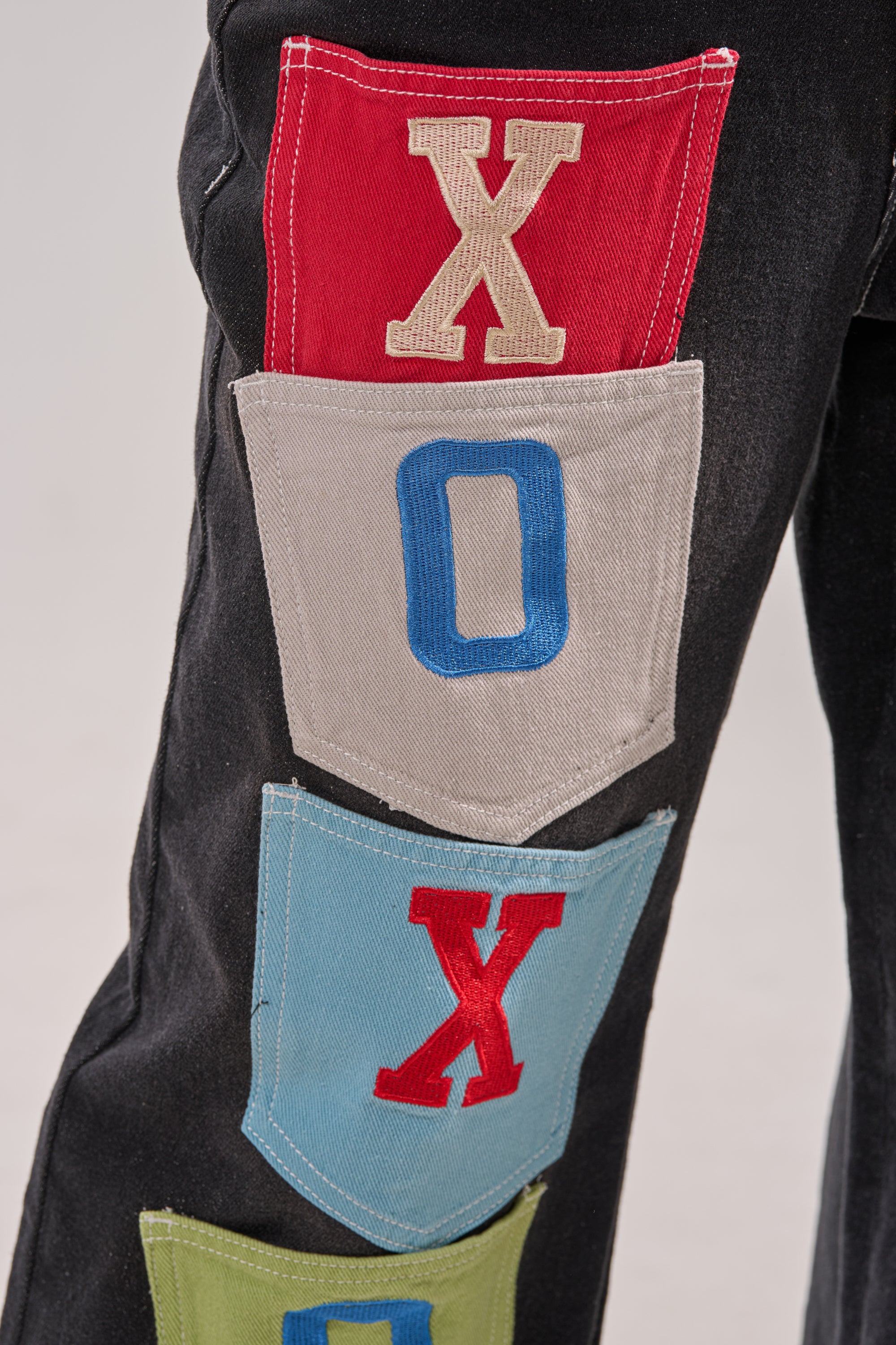 A close-up of the BRING IT BACK DENIM PATCHWORK PANT: black wide-leg jeans with colorful patch pockets, each boldly embroidered—a red "X" on red, a blue "O" on gray, and another red "X" on blue. 8