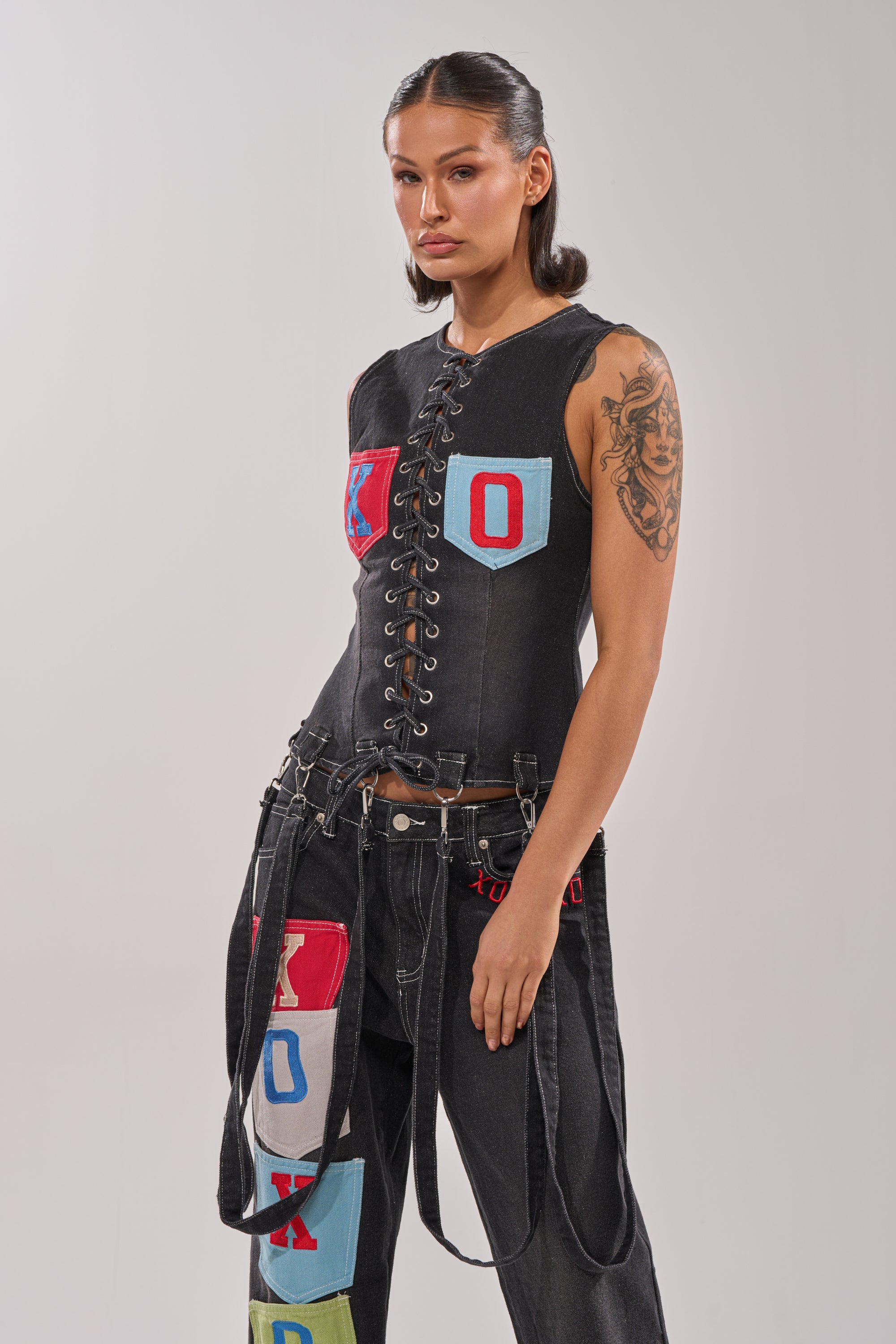 A woman with slicked-back hair models the BRING IT BACK LACE UP DENIM TOP in black-wash with a lace-up front and colorful patches, paired with black jeans featuring letter patches and straps. She reveals a tattoo on her left arm. 0