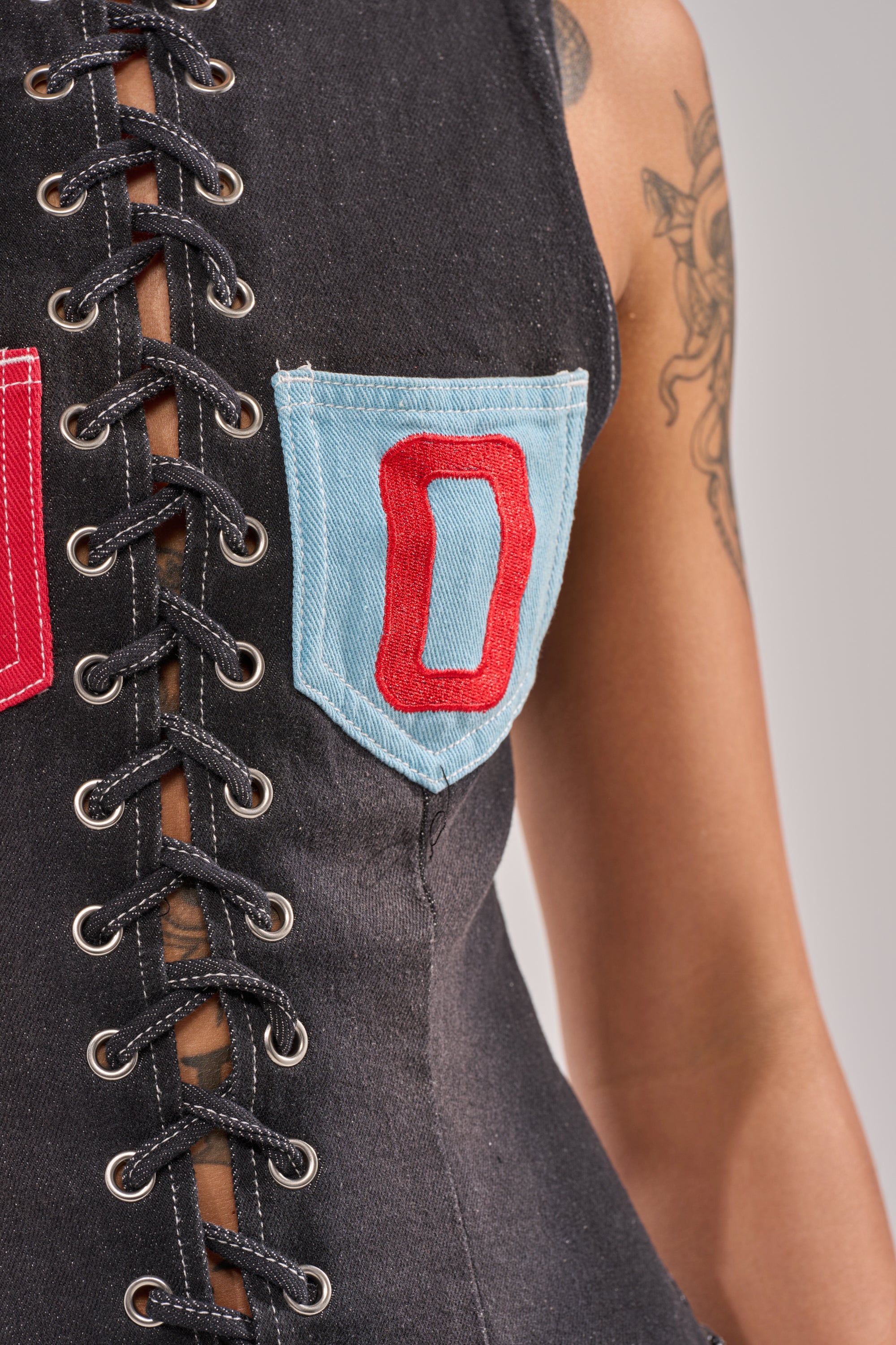 Close-up of the BRING IT BACK LACE UP DENIM TOP featuring a black-wash finish, corset-style lace-up front, bold red "O" patch pockets in red and blue, and a partial glimpse of a tattooed arm. 9