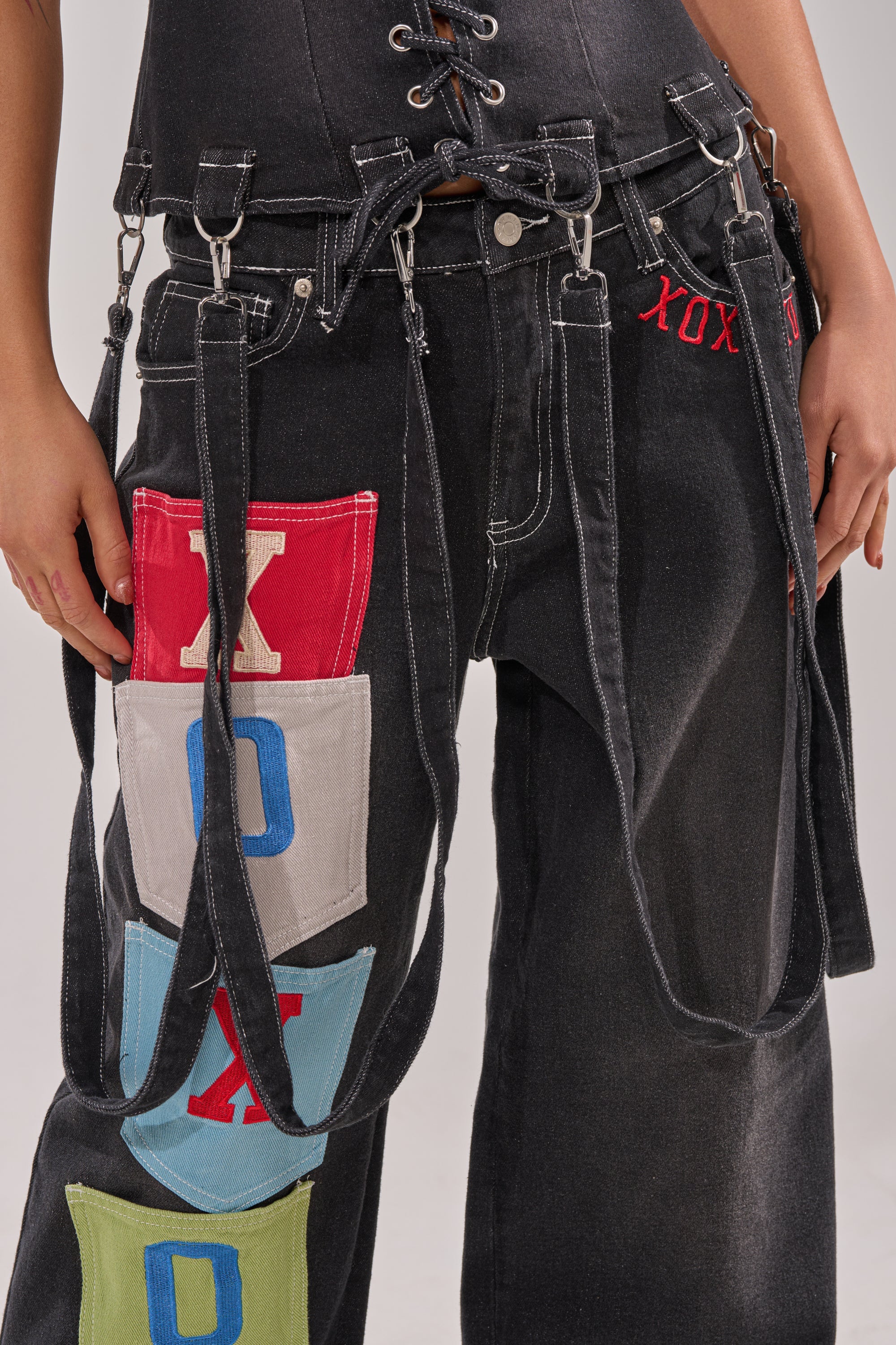 Someone wears black jeans with colorful letter patches, black straps, and metal buckles, paired with the BRING IT BACK LACE UP DENIM TOP—a black-wash corset-style denim piece featuring bold red "XOX" embroidery at the waistband. 10