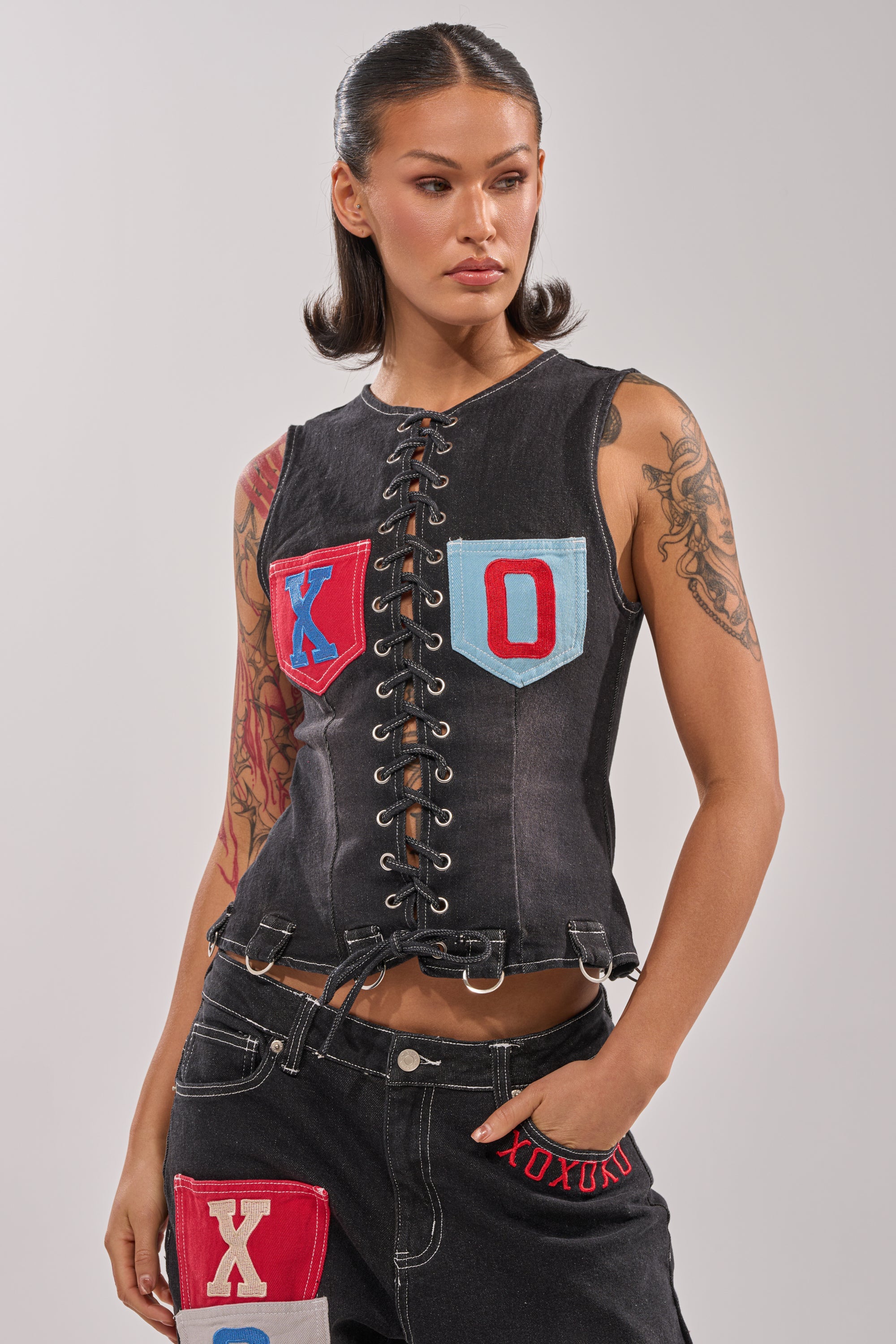 A woman with tattooed arms wears the BRING IT BACK LACE UP DENIM TOP, a corset-inspired black lace-up style with red and blue "X" and "O" patches, paired with black pants featuring "XOXO" text, patches, and detachable garter straps. 1