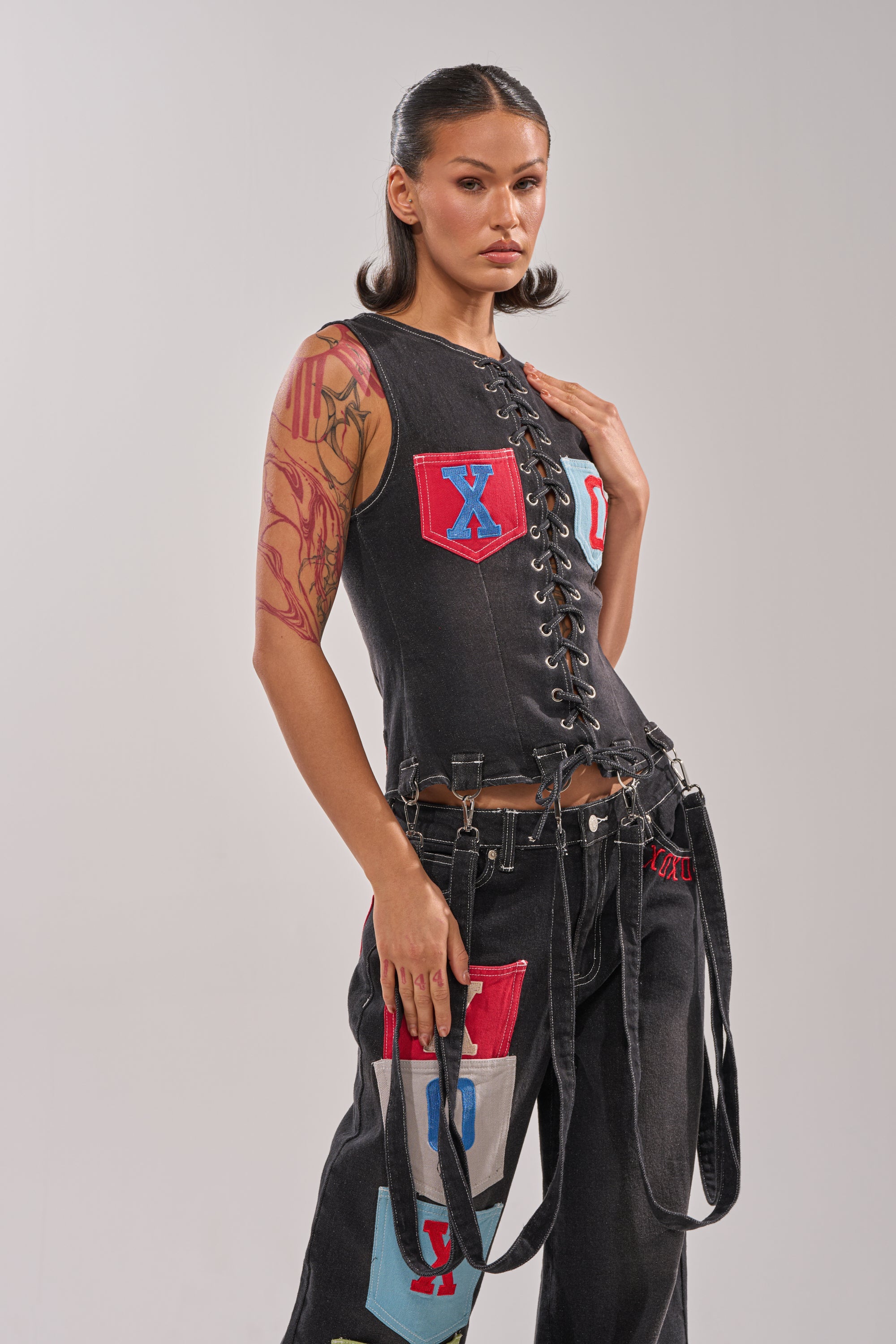 A woman with dark hair styled back wears the BRING IT BACK LACE UP DENIM TOP, a corset-inspired black lace-up piece with red and blue patches, paired with matching black pants and detachable garter straps, posing confidently against a plain background. 2