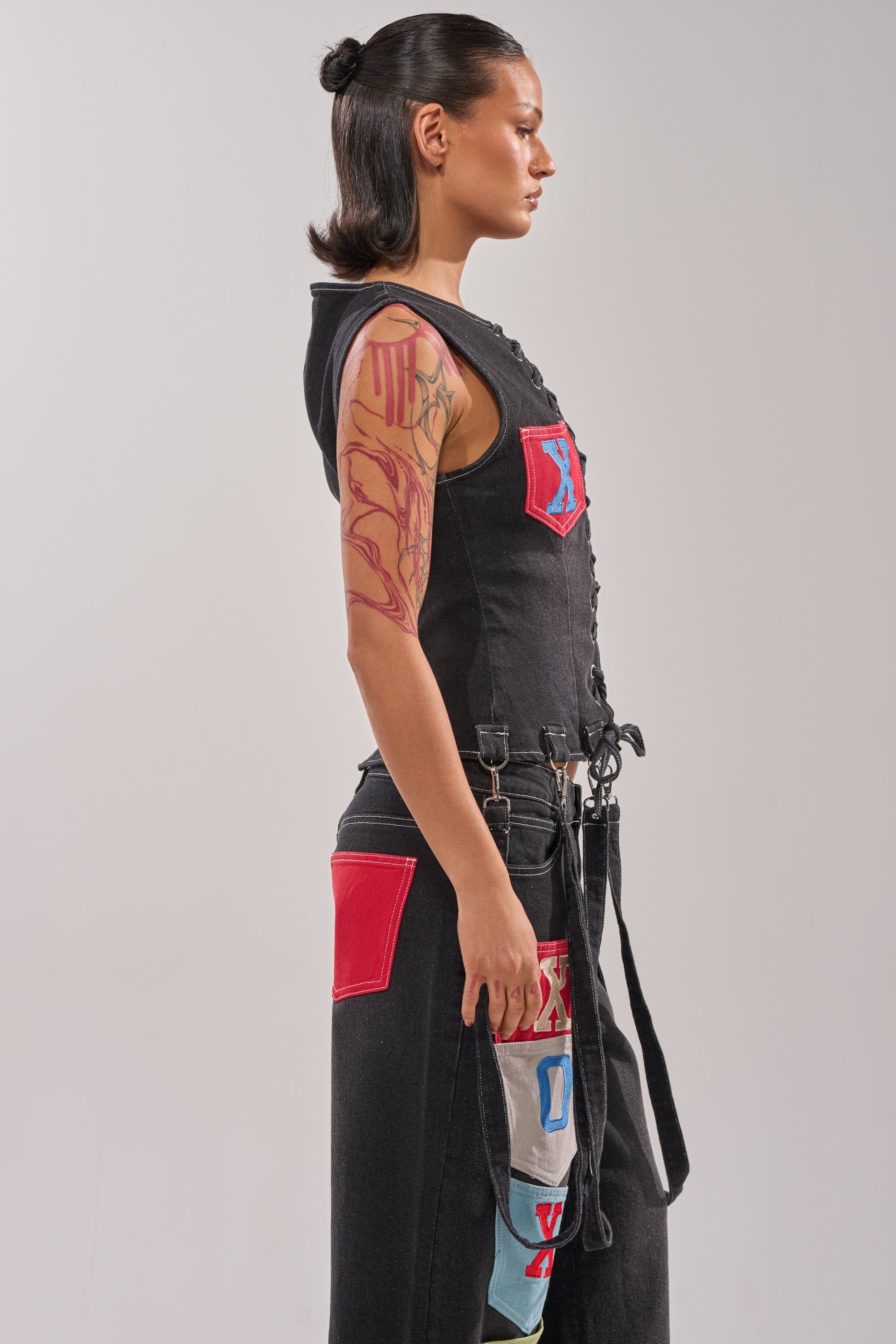 A woman in profile against a plain background wears the BRING IT BACK LACE UP DENIM TOP, featuring a corset-inspired silhouette with colorful denim patches, lace-up front, detachable garter straps, sleek bun, and a red upper arm tattoo. 3