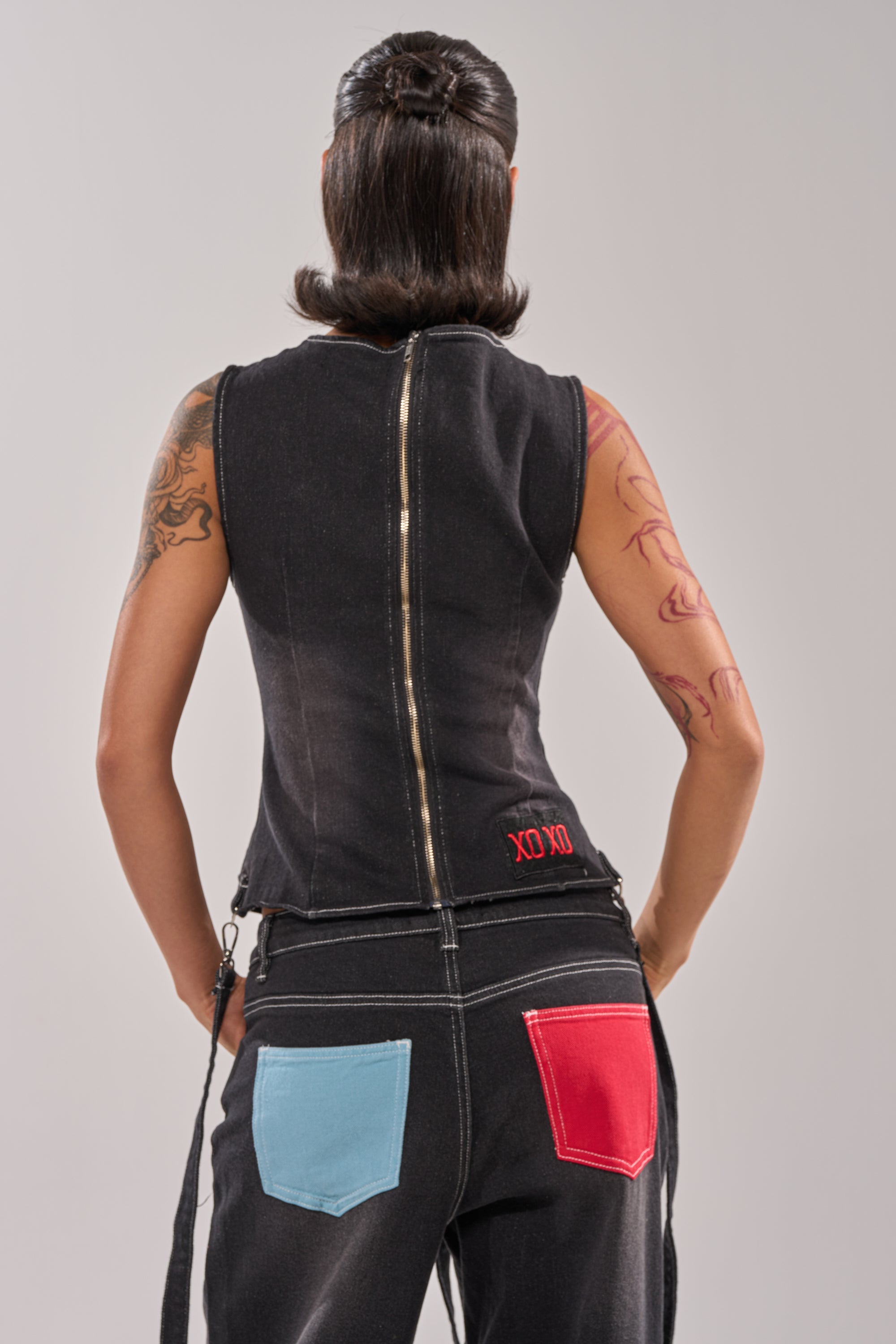 A person with dark, styled hair faces away, wearing the BRING IT BACK LACE UP DENIM TOP in black-wash denim with a lace-up front and zip back, paired with black pants featuring blue and red pockets. Visible tattoos adorn their arms. 4