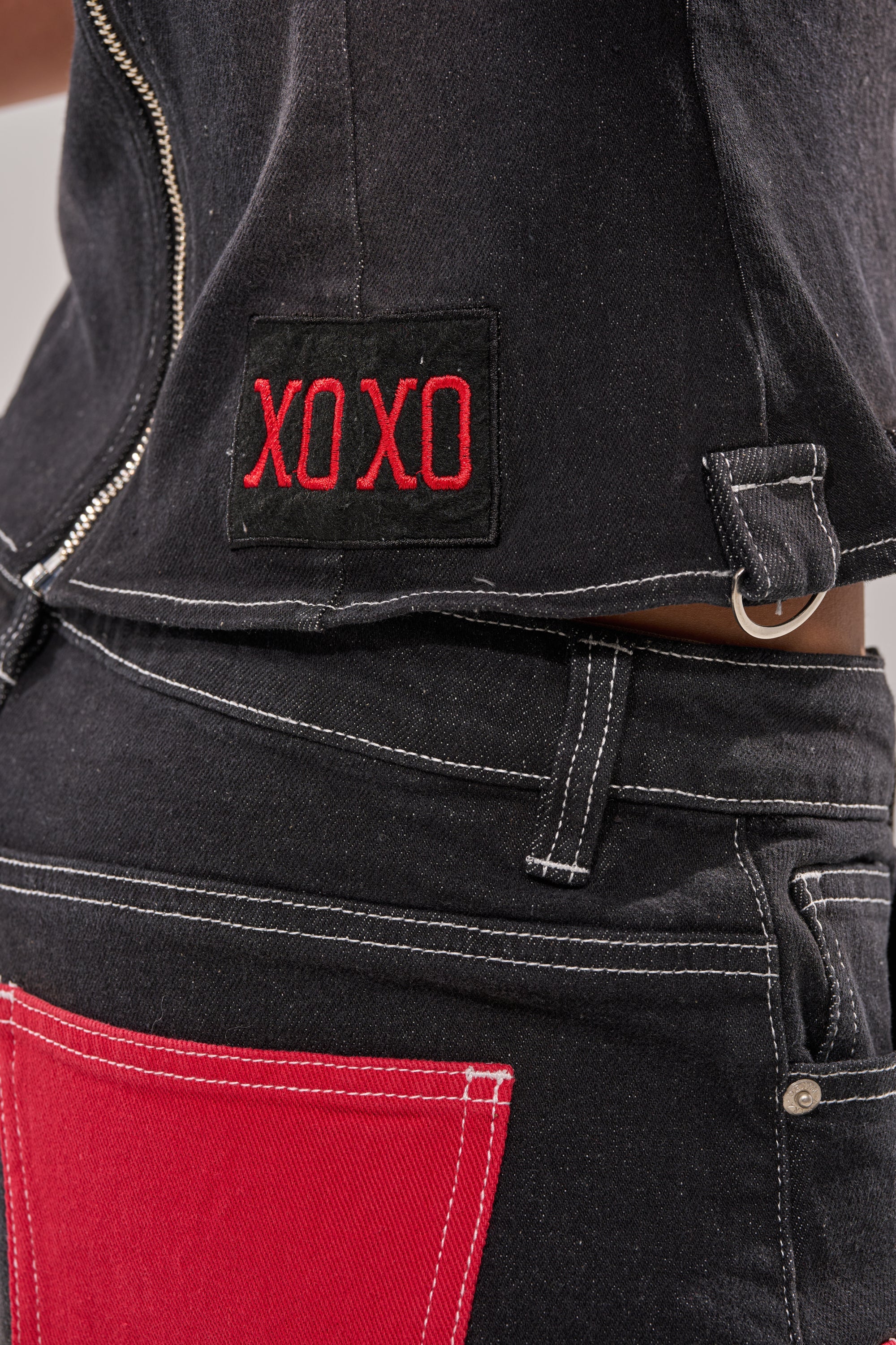 Close-up of a person in the BRING IT BACK LACE UP DENIM TOP, a black-wash denim piece with white stitching, corset-inspired fit, front zipper, red "XO XO" patch, and paired with pants featuring a striking bright red back pocket. 8