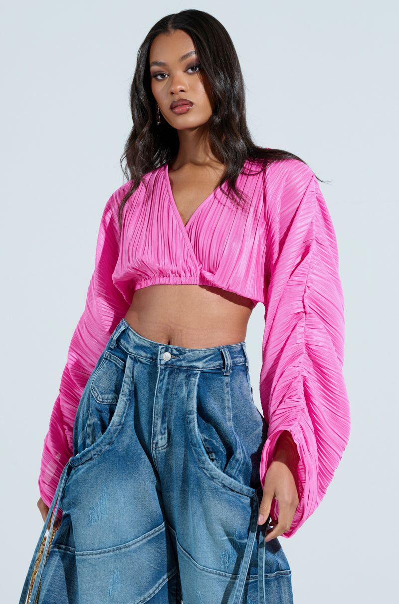 A woman poses confidently against a plain backdrop, wearing a vibrant pink Cropped Plisse Ruched Sleeve Blouse paired with loose-fitting, high-waisted denim jeans featuring decorative stitching.