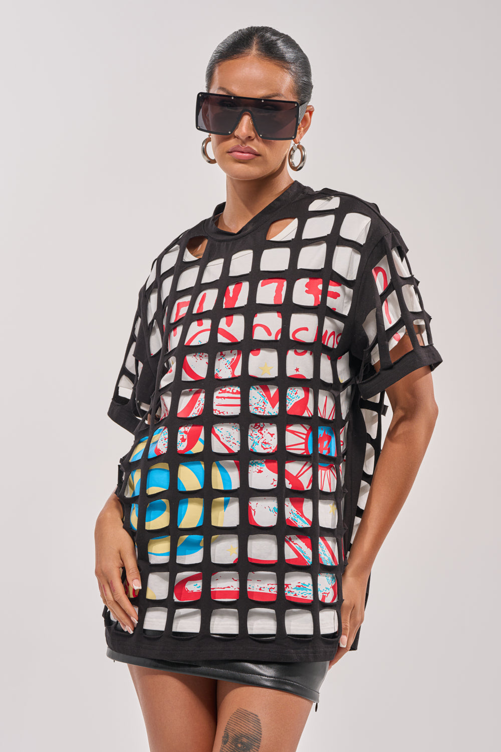 Wearing large black sunglasses and hoop earrings, a woman confidently poses in the CHECKED OUT OVERSIZED TEE layered under a black mesh top with a black leather mini skirt, creating a bold streetwear look against a plain background. 0