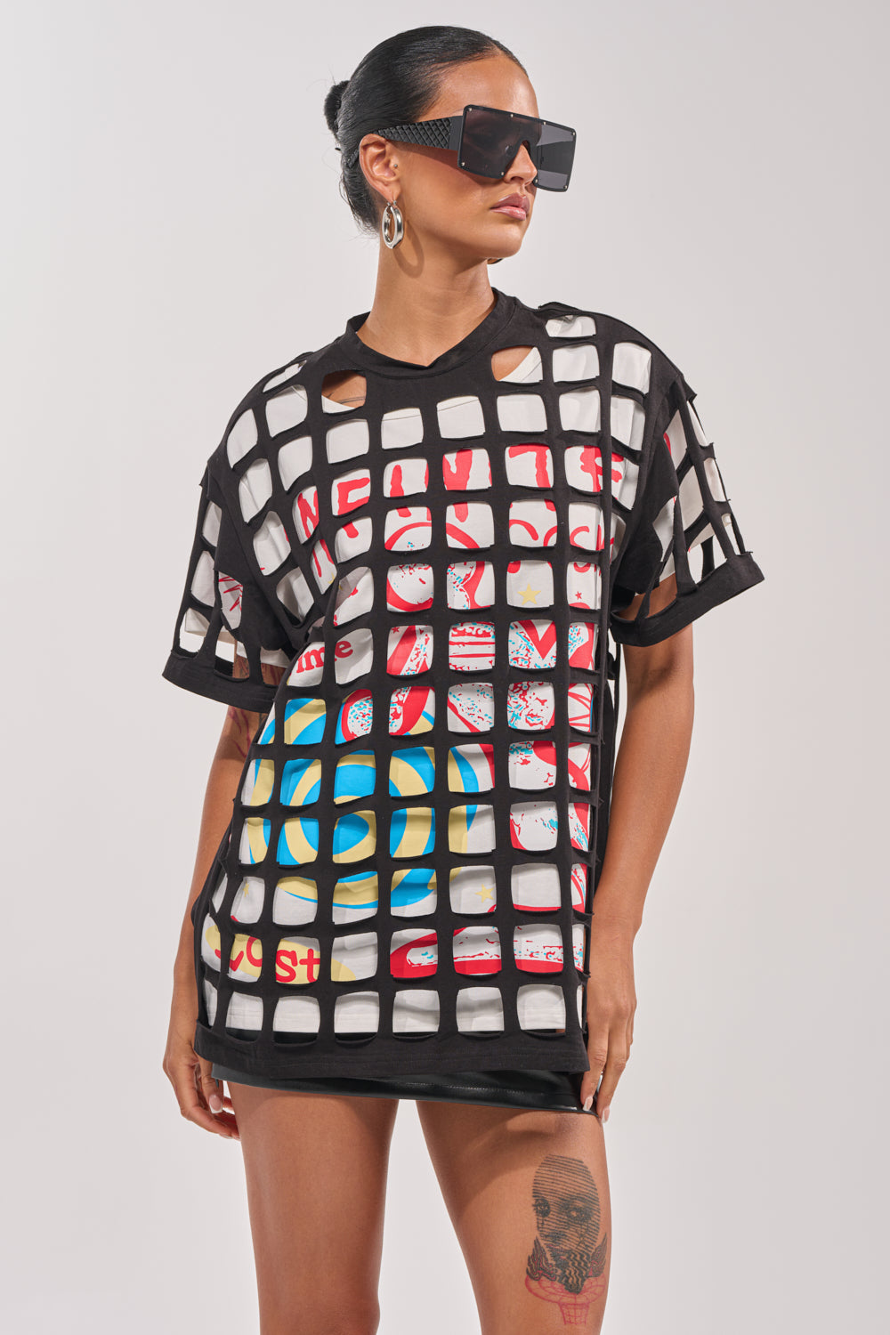 A woman exudes confidence in the CHECKED OUT OVERSIZED TEE—styled over a bold graphic top with black sunglasses and her hair tied back—highlighting her thigh tattoo. A perfect choice for making a statement at any festival. 1