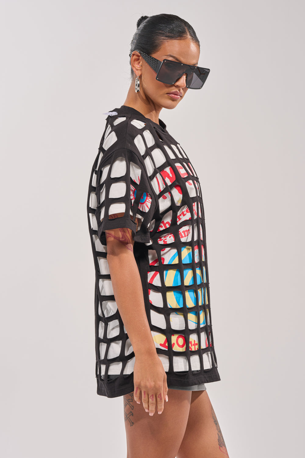 A woman in streetwear style stands sideways against a plain light background, wearing oversized black square sunglasses, a black mesh t-shirt layered over the CHECKED OUT OVERSIZED TEE, with her hair tied back. 2