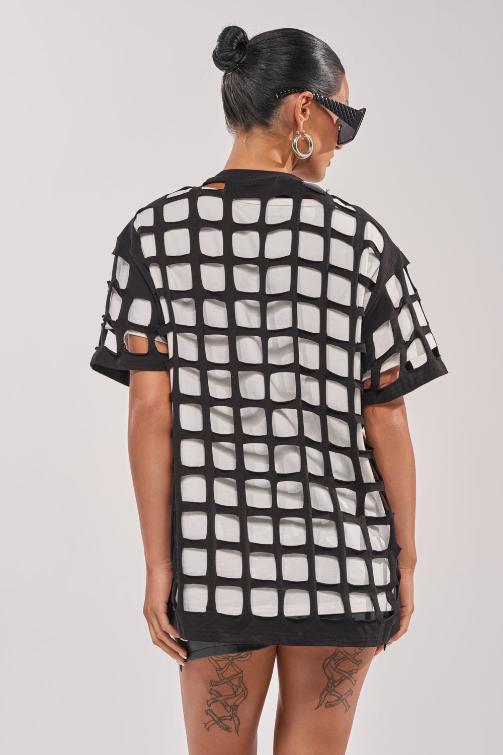A woman with a bun and sunglasses wears the CHECKED OUT OVERSIZED TEE, a black short-sleeve graphic top with a bold white square grid cutout pattern, revealing her back and thigh tattoos—perfect for streetwear or festival style. 3