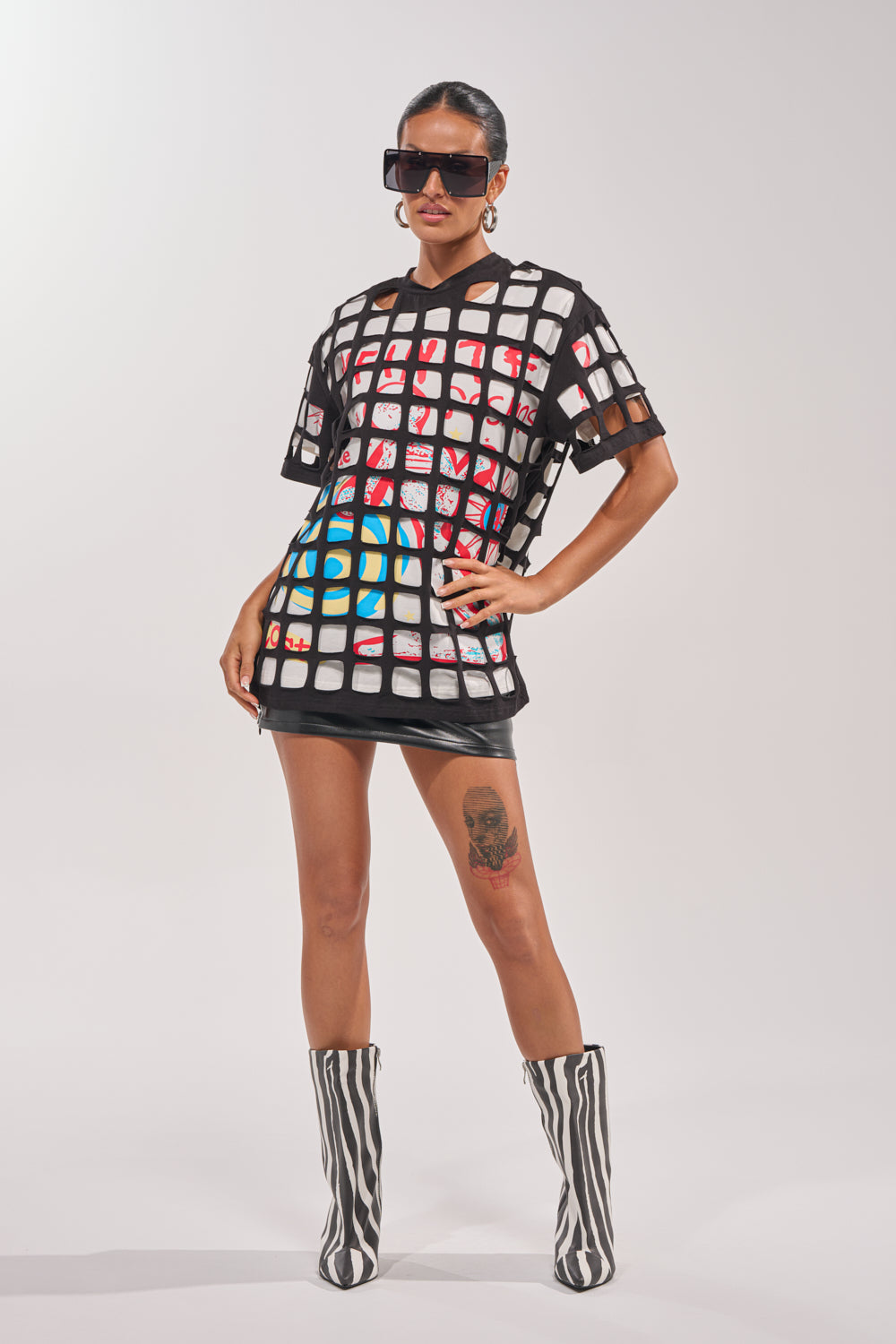 A woman rocks the CHECKED OUT OVERSIZED TEE as a see-through grid dress over a colorful layer, styling it with oversized sunglasses, hoop earrings, zebra-striped knee-high boots, and showing off a thigh tattoo—perfect for festival fashion. 4