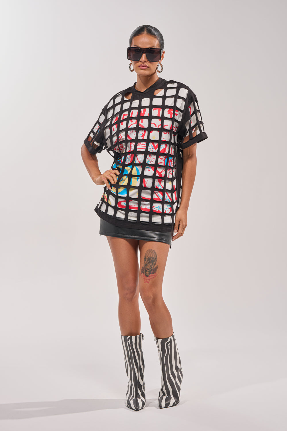 A person exudes streetwear vibes in the CHECKED OUT OVERSIZED TEE layered over a colorful shirt, black mini skirt, striped boots, oversized sunglasses, hoops, and a thigh tattoo—posing confidently against a plain light background. 5