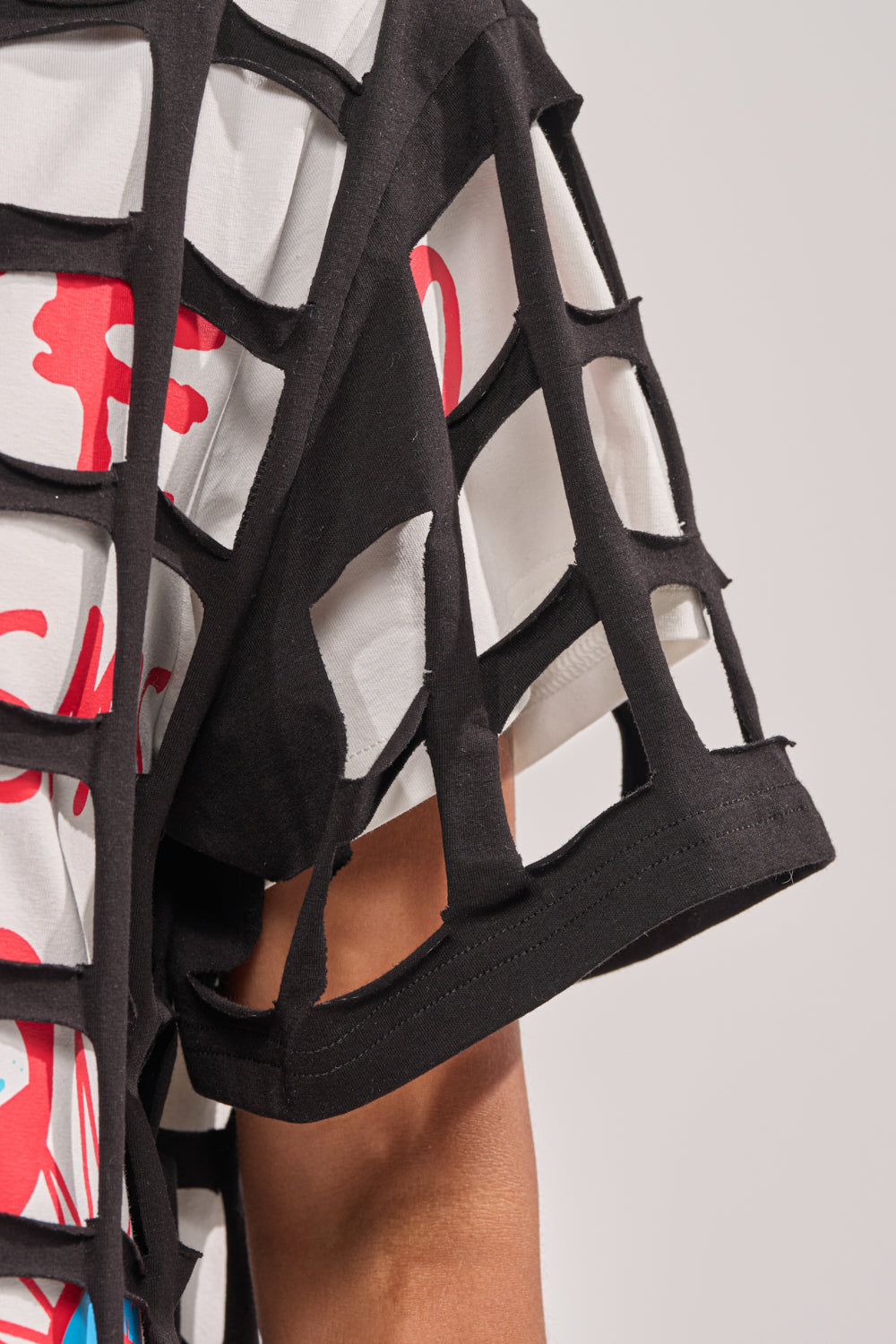 A close-up of a person's arm and shoulder wearing the CHECKED OUT OVERSIZED TEE, featuring a bold red and blue graphic and layered with a black, grid-patterned cut-out mesh—ideal for festival standout style. 6