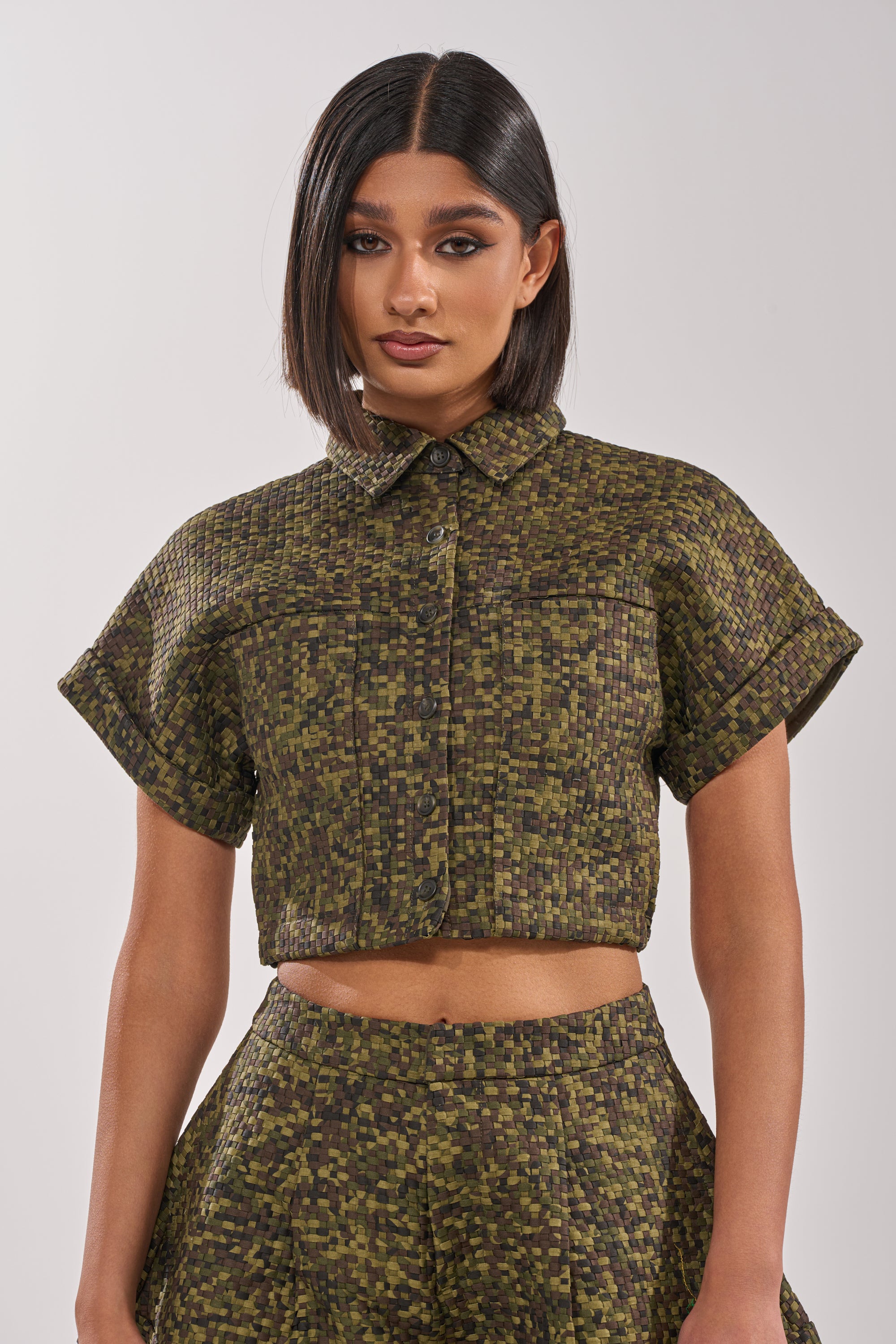 A woman with straight, shoulder-length dark hair wears the DOWN UNDER BUTTON DOWN, a cropped, short-sleeved button-up shirt with matching high-waisted shorts in a green and brown pixelated pattern, standing against a plain background. 0