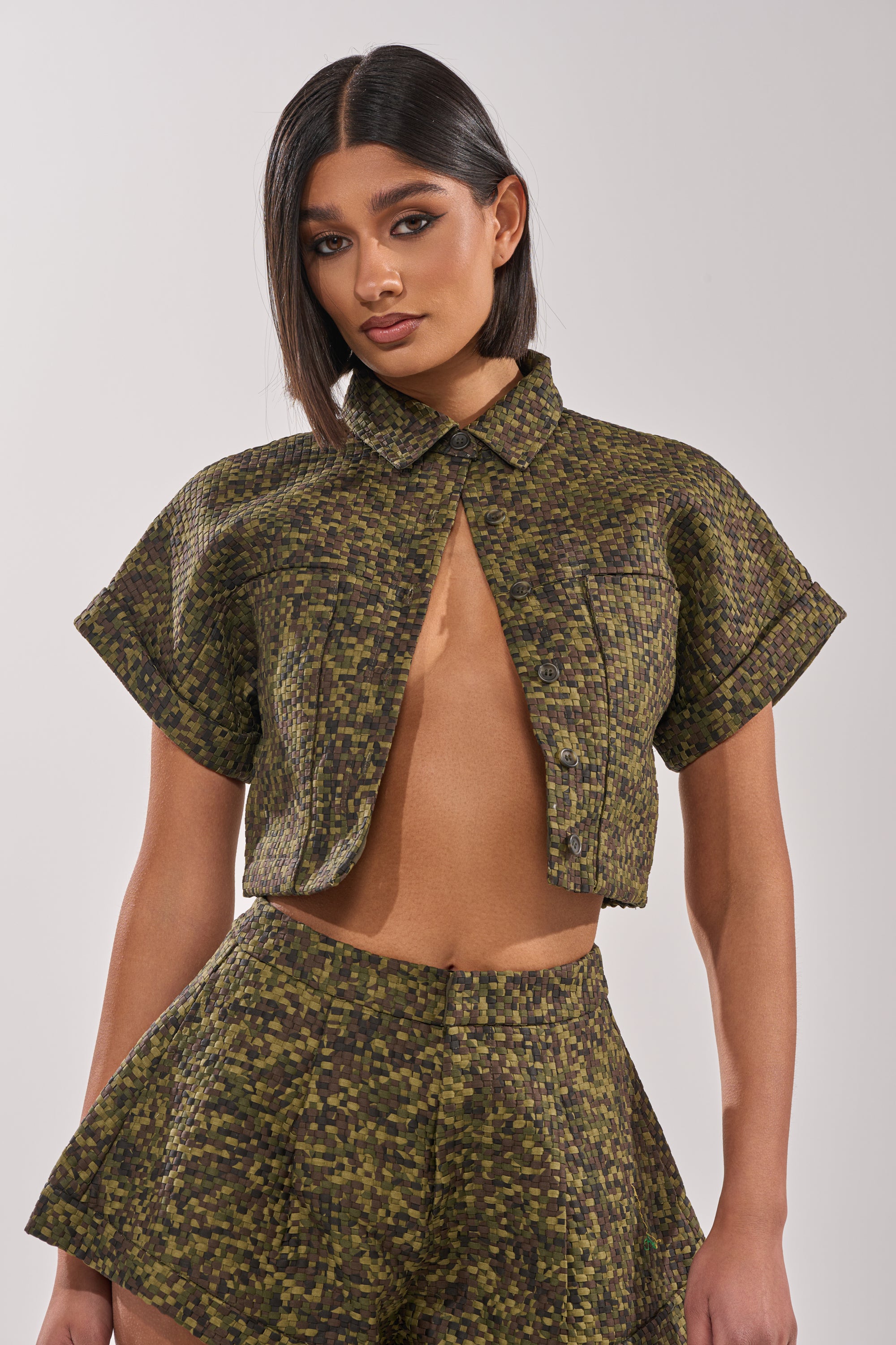 A woman with straight dark hair models the DOWN UNDER BUTTON DOWN, a green and brown textured cropped jacket with short sleeves and matching high-waisted shorts, posing against a plain light background. 1