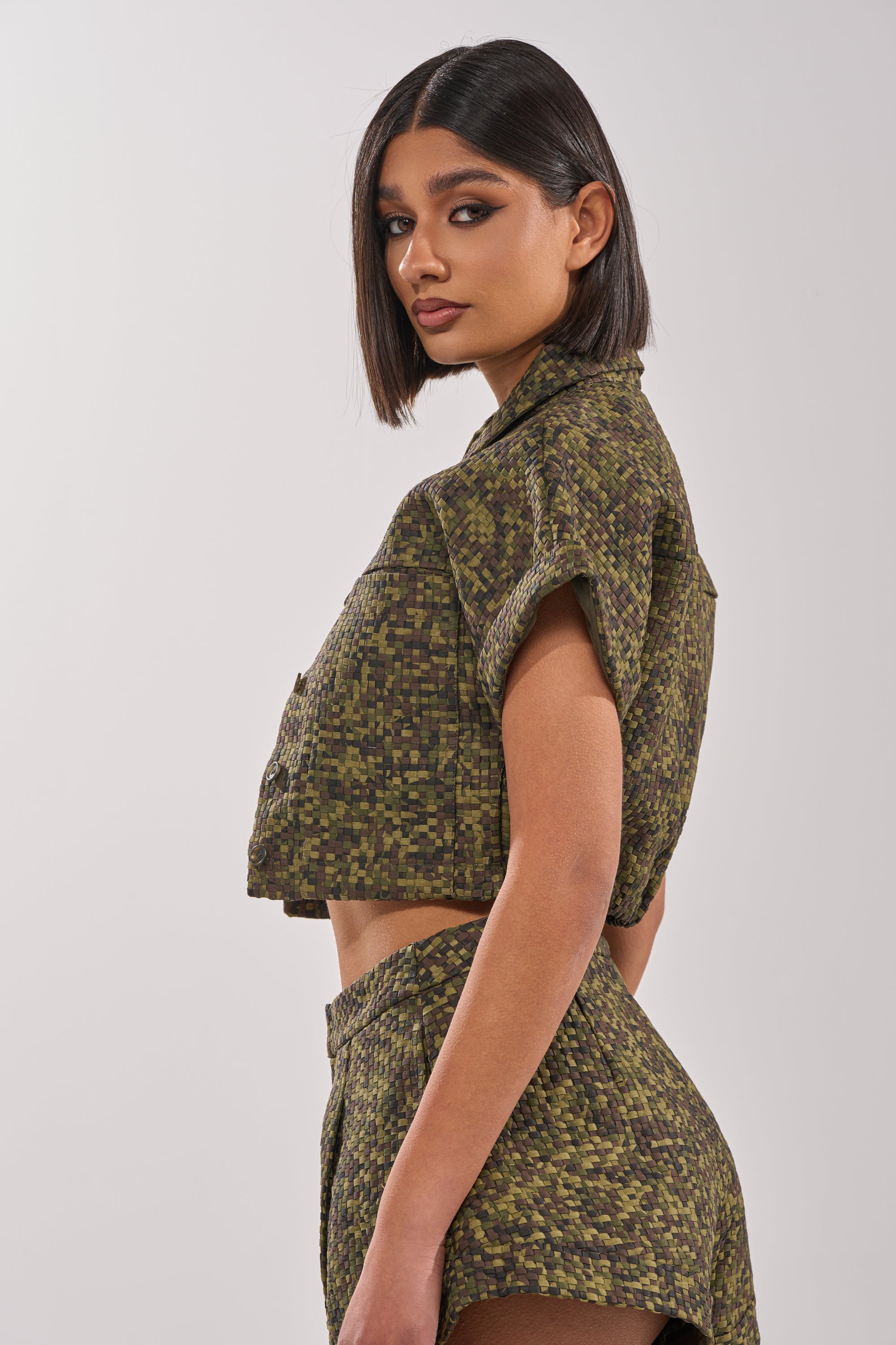 A woman with straight, shoulder-length dark hair models the DOWN UNDER BUTTON DOWN top and matching high-waisted shorts in rattan textured fabric, standing sideways and glancing over her shoulder against a plain light background. 3