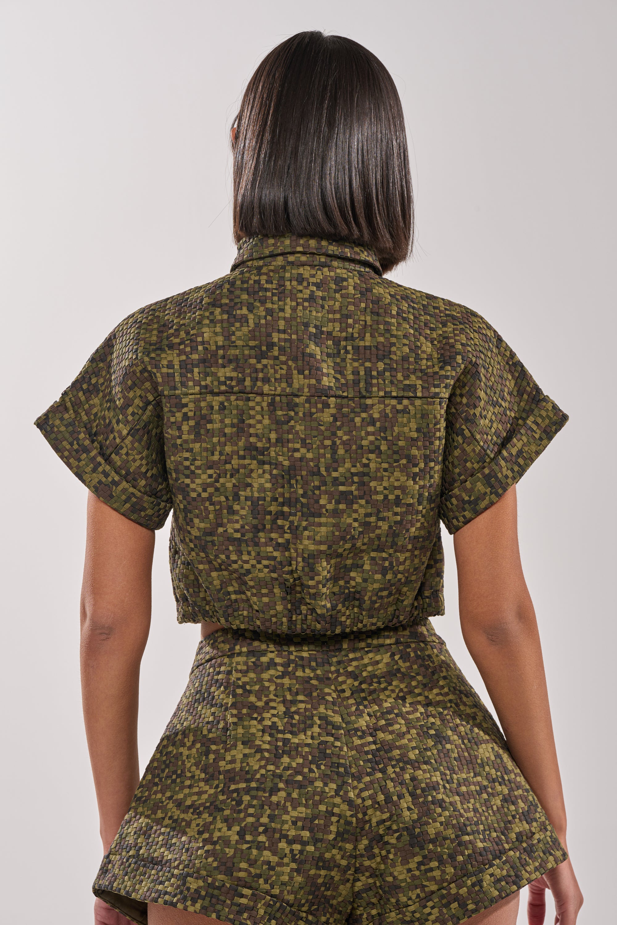 A person with straight, shoulder-length dark hair is seen from behind wearing the DOWN UNDER BUTTON DOWN, a cropped boxy blouse with matching high-waisted shorts in a green and brown pixelated print. The background is plain and light-colored. 4