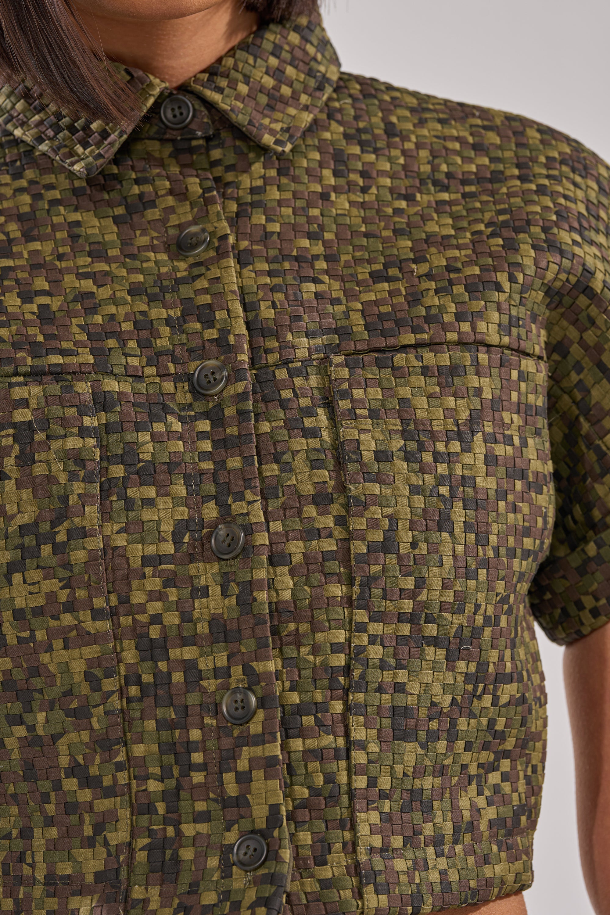 A close-up of a person wearing the DOWN UNDER BUTTON DOWN—a textured, olive green, short-sleeve, cropped shirt with a woven green, brown, and black checkered pattern, collar, and dark front buttons. 8