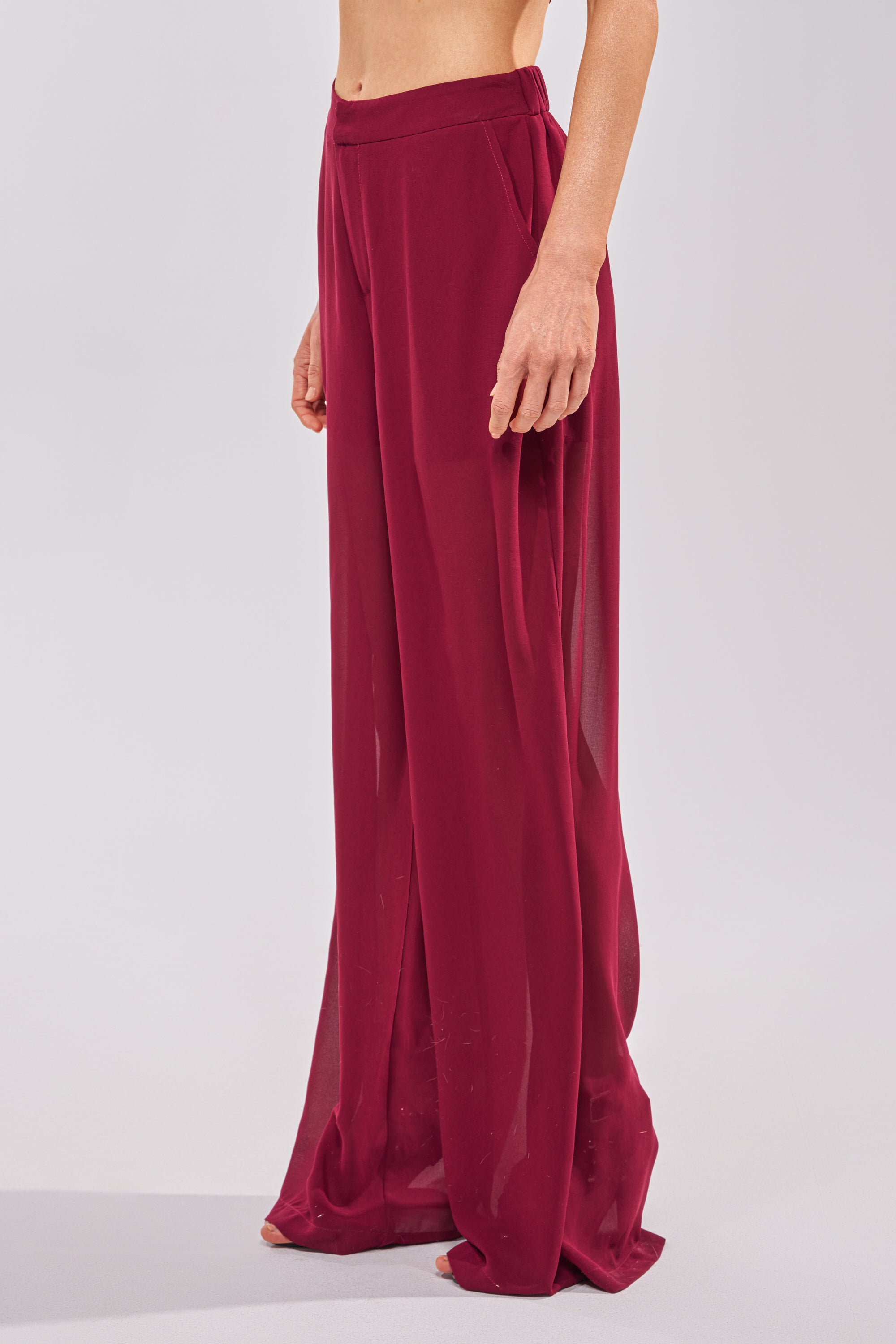 A person models the COOL GIRL TROUSER—high-rise, sheer burgundy chiffon pants—barefoot against a plain light background, showing only their lower torso, legs, and one arm. 1