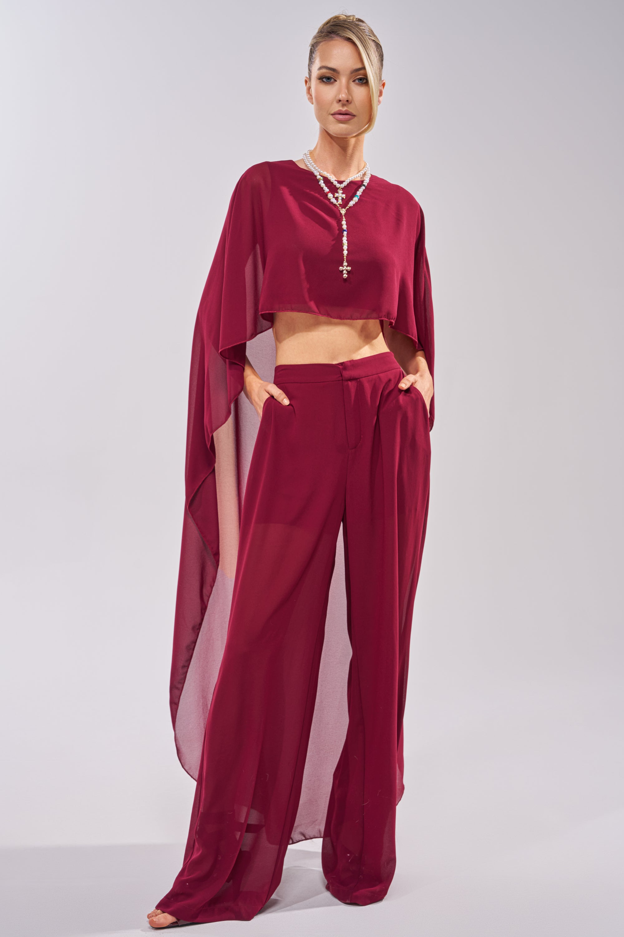 A woman wears the COOL GIRL TROUSER, a matching burgundy set featuring chiffon wide-leg pants and a cape-style top, accessorized with layered necklaces and hands in her pockets, posed against a light background. 3