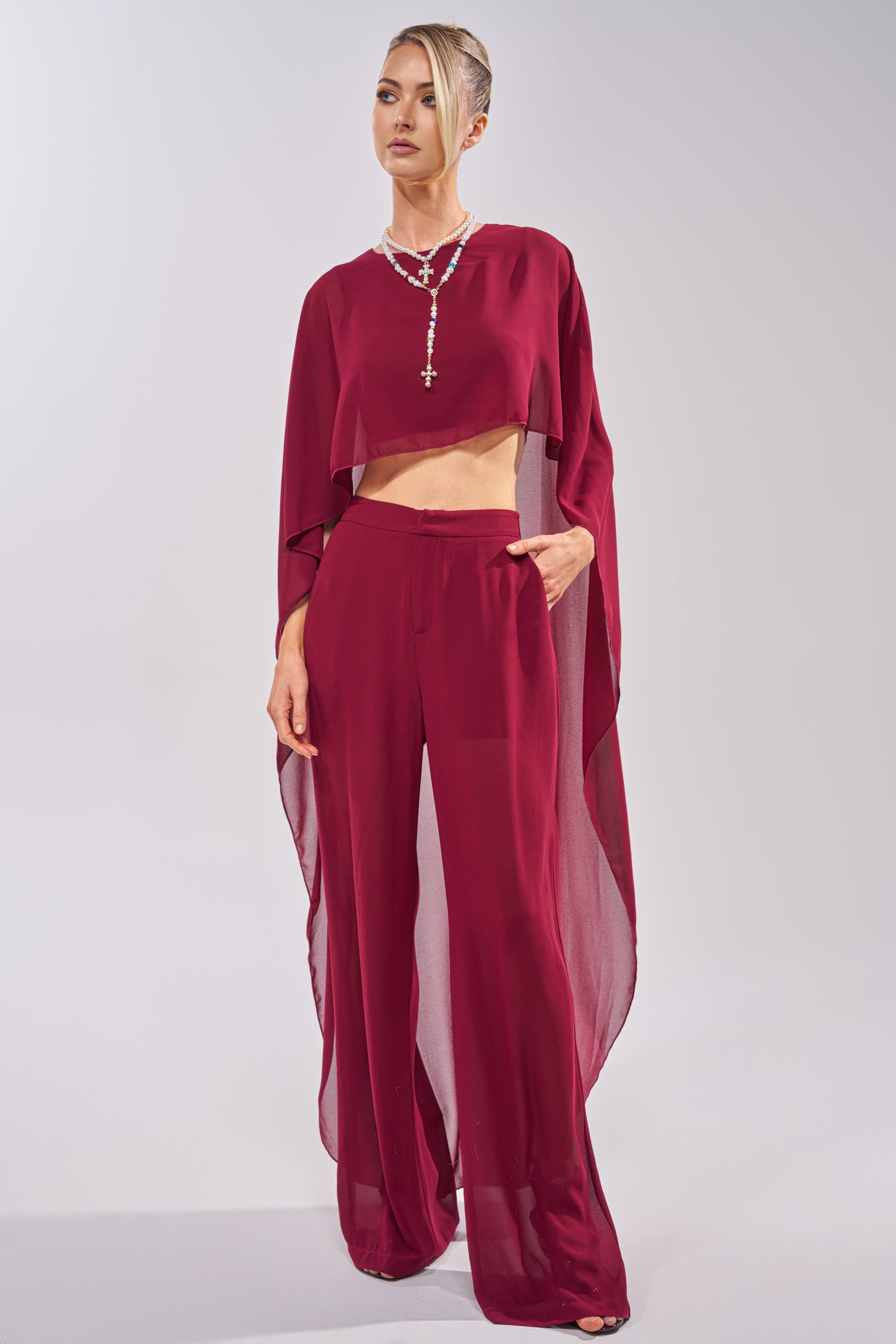 A woman poses against a neutral background in the COOL GIRL TROUSER—a stylish, sheer burgundy high-rise chiffon pant—paired with a loose, flowing asymmetrical top and beaded necklace; her hair is styled up and one hand is in her pocket. 5