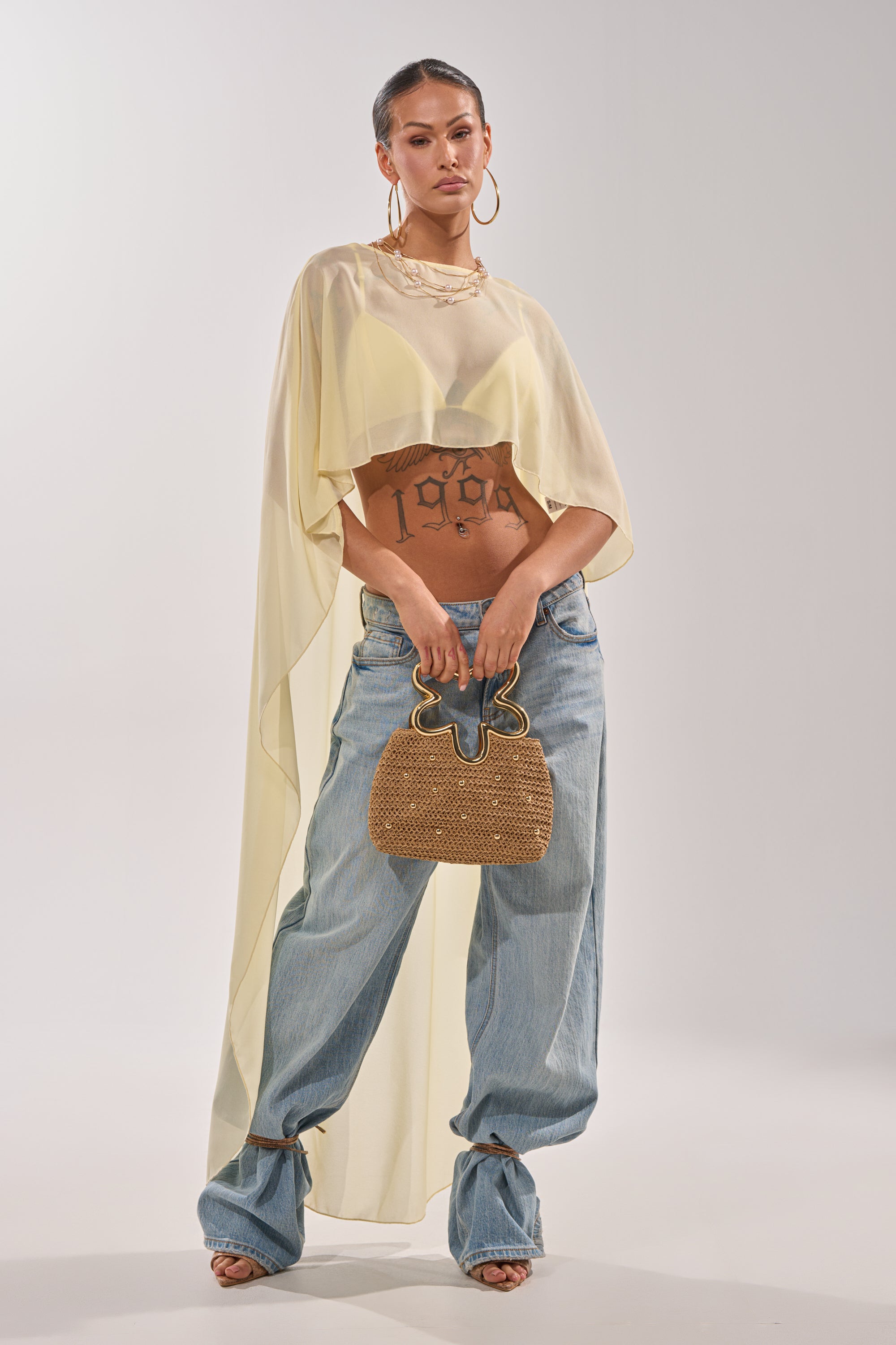 A woman exudes confidence in the COOL GIRL CHIFFON BLOUSE IN YELLOW, styled over a crop top with loose light blue jeans, open-toe heels, a woven handbag, large hoop earrings, and visible tattoos. 0