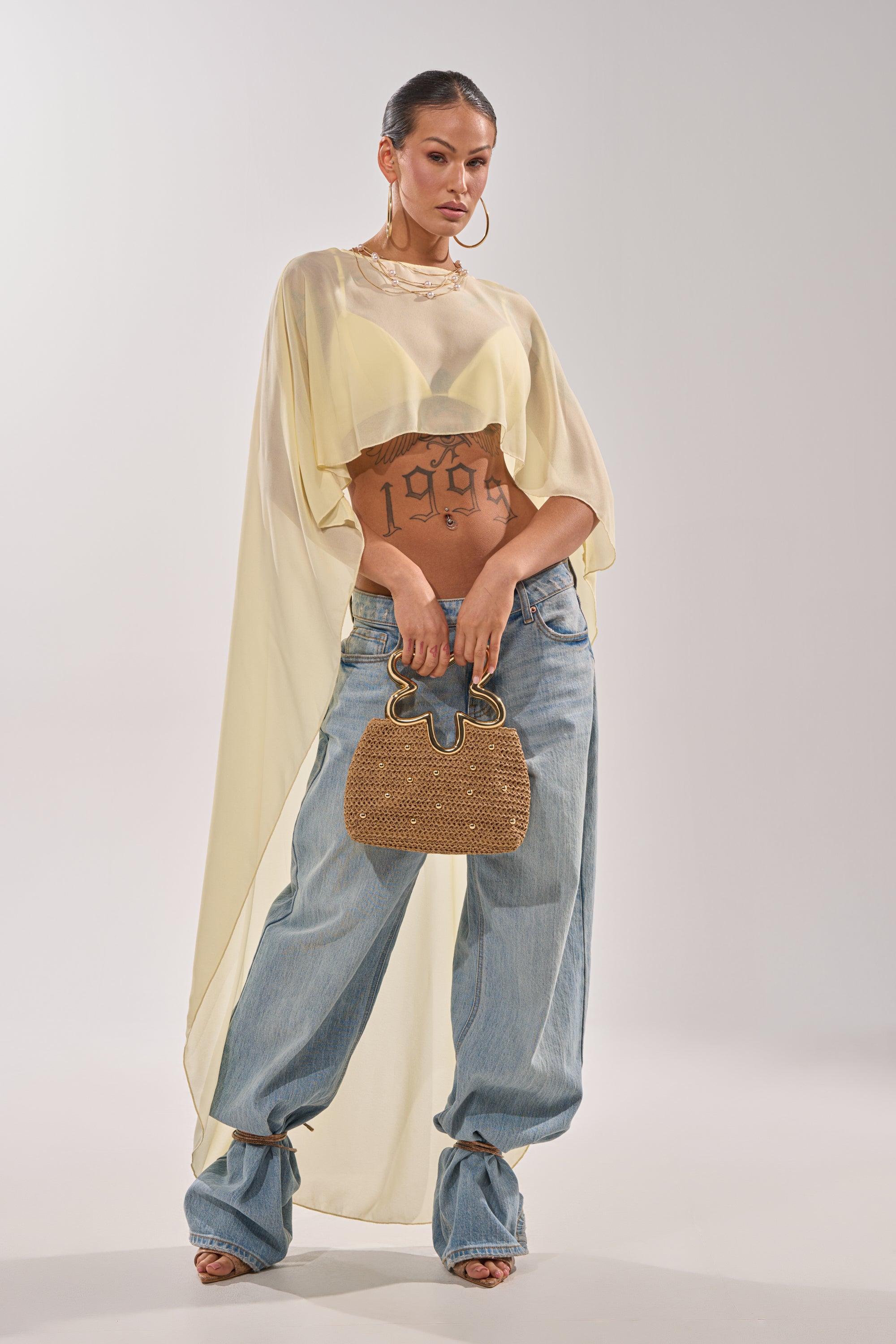 A woman exudes confidence in the COOL GIRL CHIFFON BLOUSE IN YELLOW and loose blue jeans, accessorized with a textured brown handbag and large hoop earrings. Her midriff with a "1997" tattoo shows beneath the sheer top. 2
