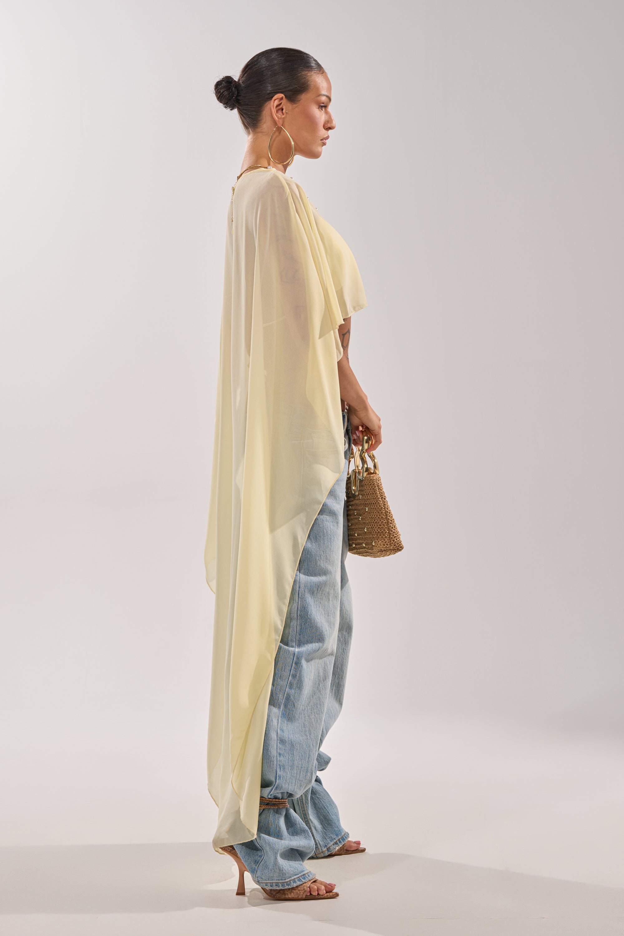 A woman in profile wears the COOL GIRL CHIFFON BLOUSE IN YELLOW over a white tank, paired with loose blue jeans, strappy heels, and a woven handbag. Her hair is neatly styled in a bun with large hoop earrings as accessories. 3
