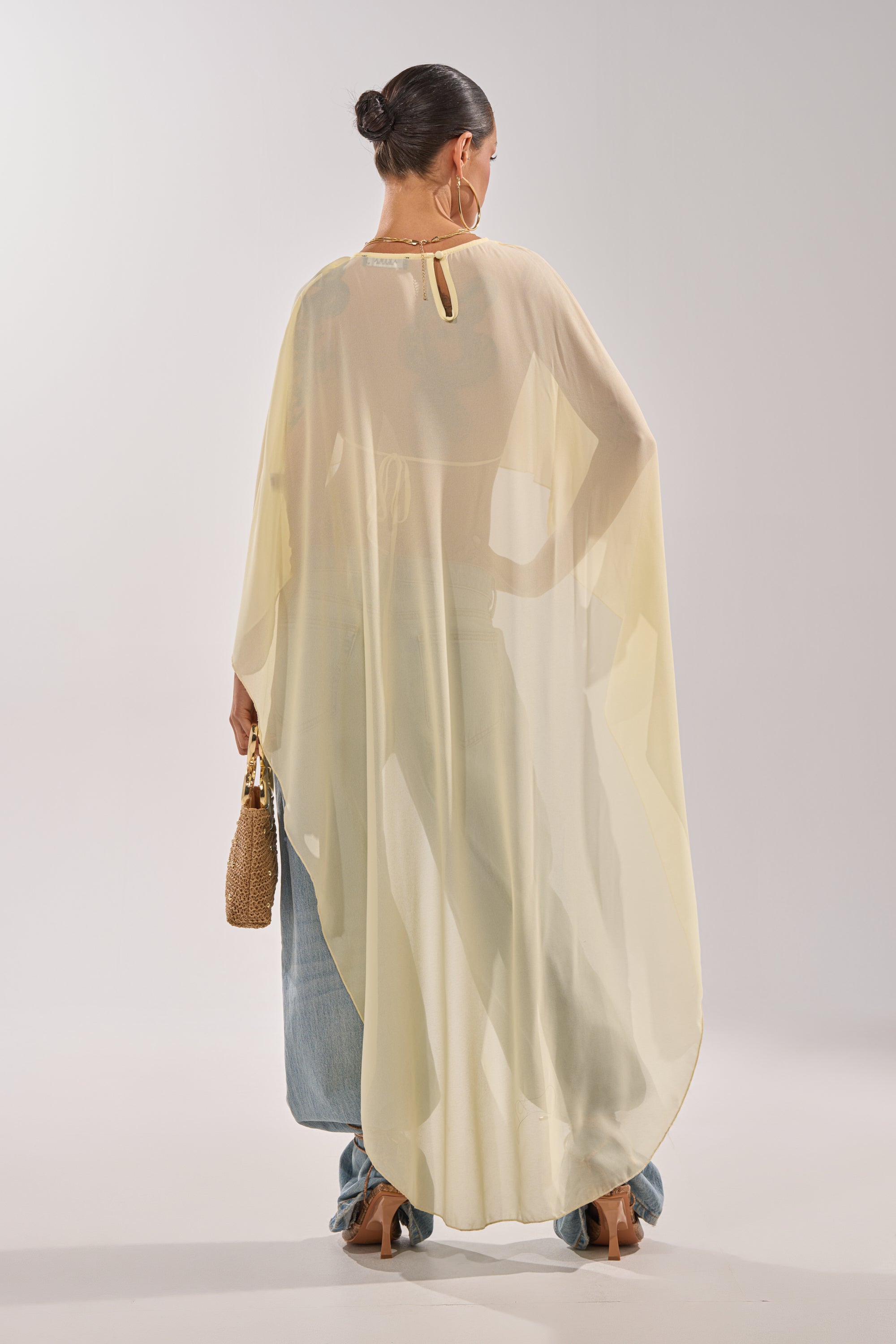A woman in a neat bun faces away, wearing the COOL GIRL CHIFFON BLOUSE IN YELLOW with light jeans and tan heels. She carries a woven handbag against a simple, light-colored background. 4