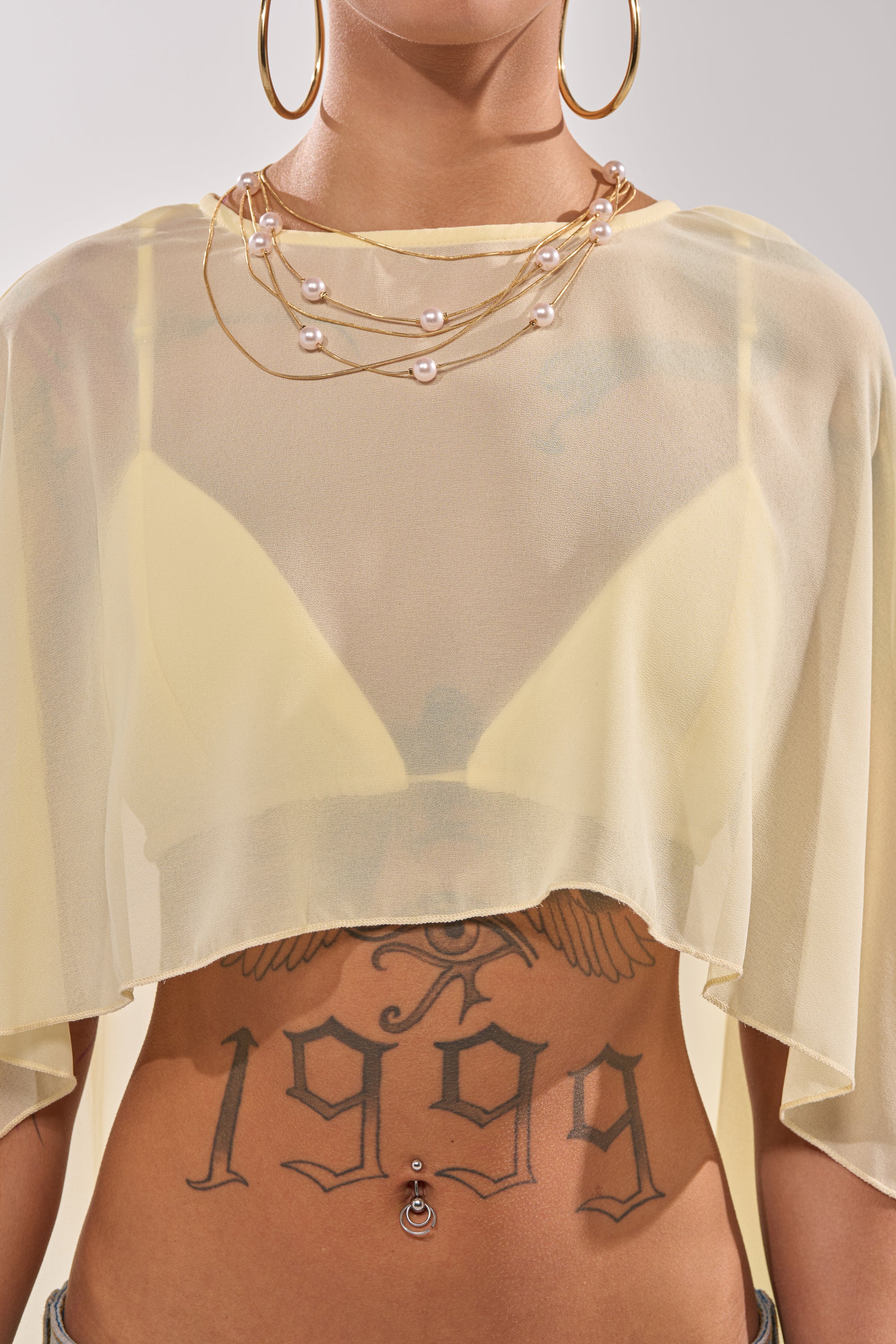 A person wears the COOL GIRL CHIFFON BLOUSE IN YELLOW over a yellow bra, styled with gold hoop earrings, layered gold and pearl necklaces, a large "1999" stomach tattoo, and a pierced belly button. 5