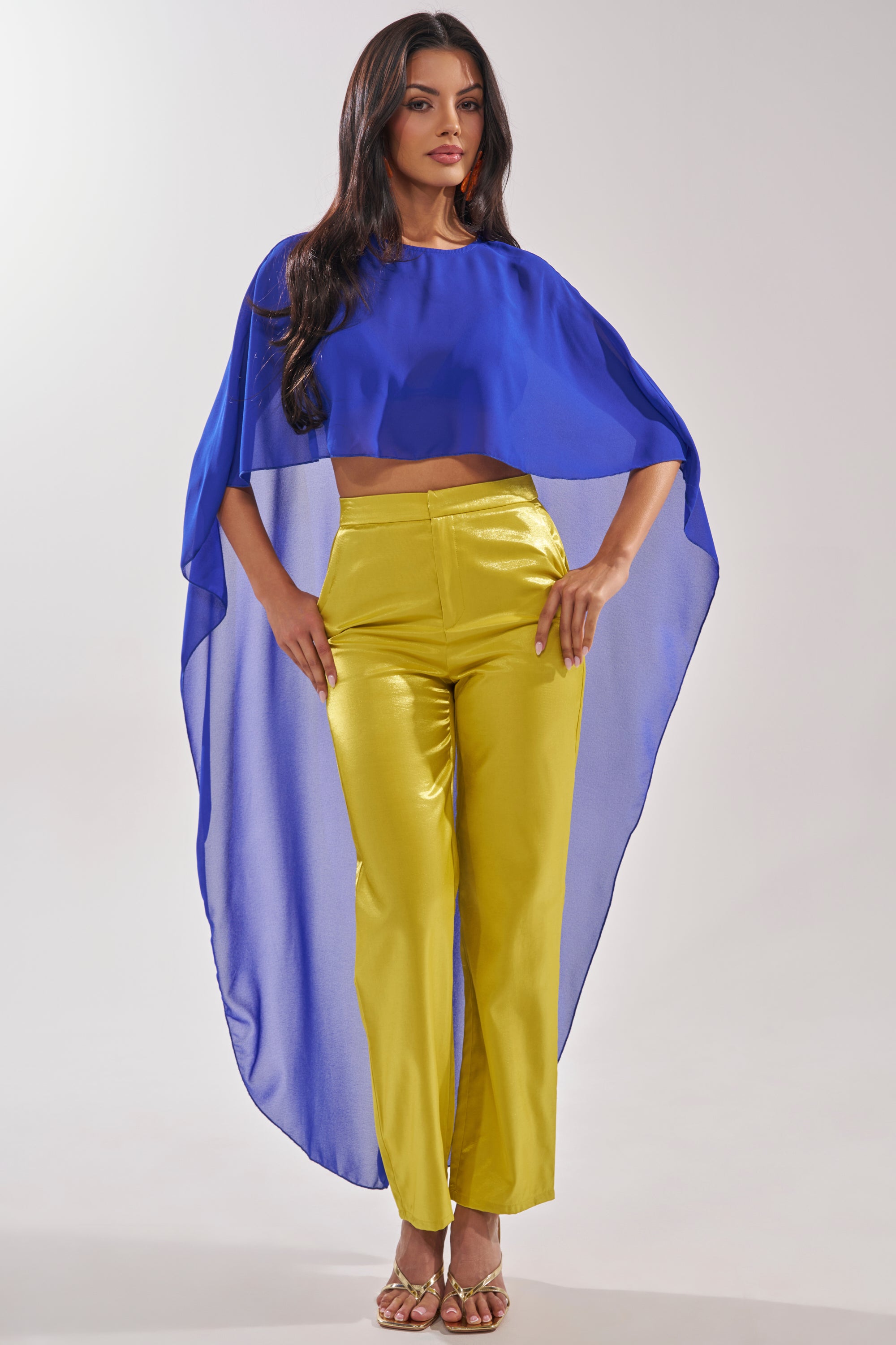 A woman stands confidently with one hand on her hip against a plain background, wearing the COOL GIRL CHIFFON BLOUSE IN ROYAL BLUE styled as a cape over a crop top and yellow pants, paired with gold sandals. 0