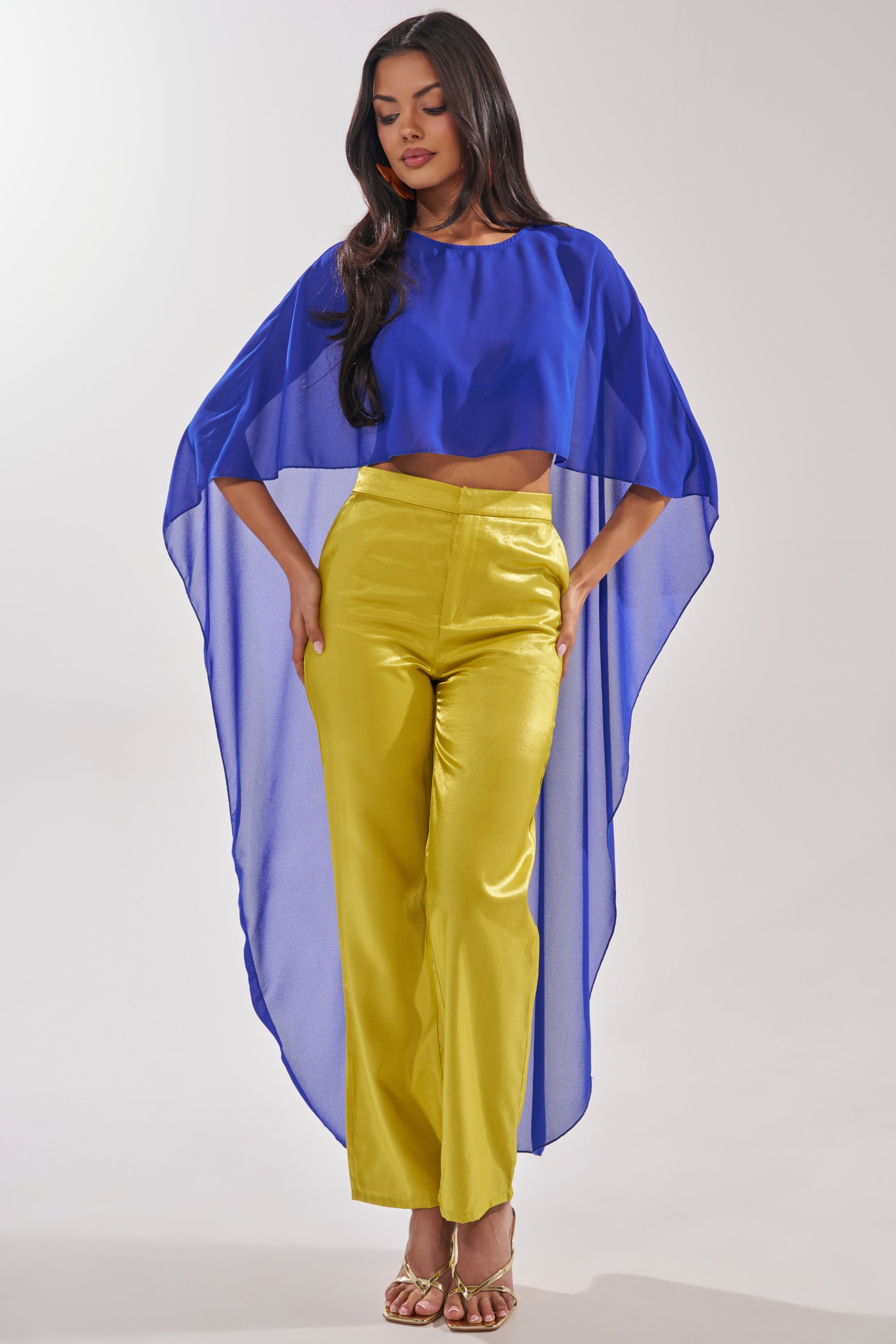 A woman models the COOL GIRL CHIFFON BLOUSE IN ROYAL BLUE, featuring sheer fabric and draped sides, paired with high-waisted yellow pants and gold heels, hands on hips as she gazes downward. 3
