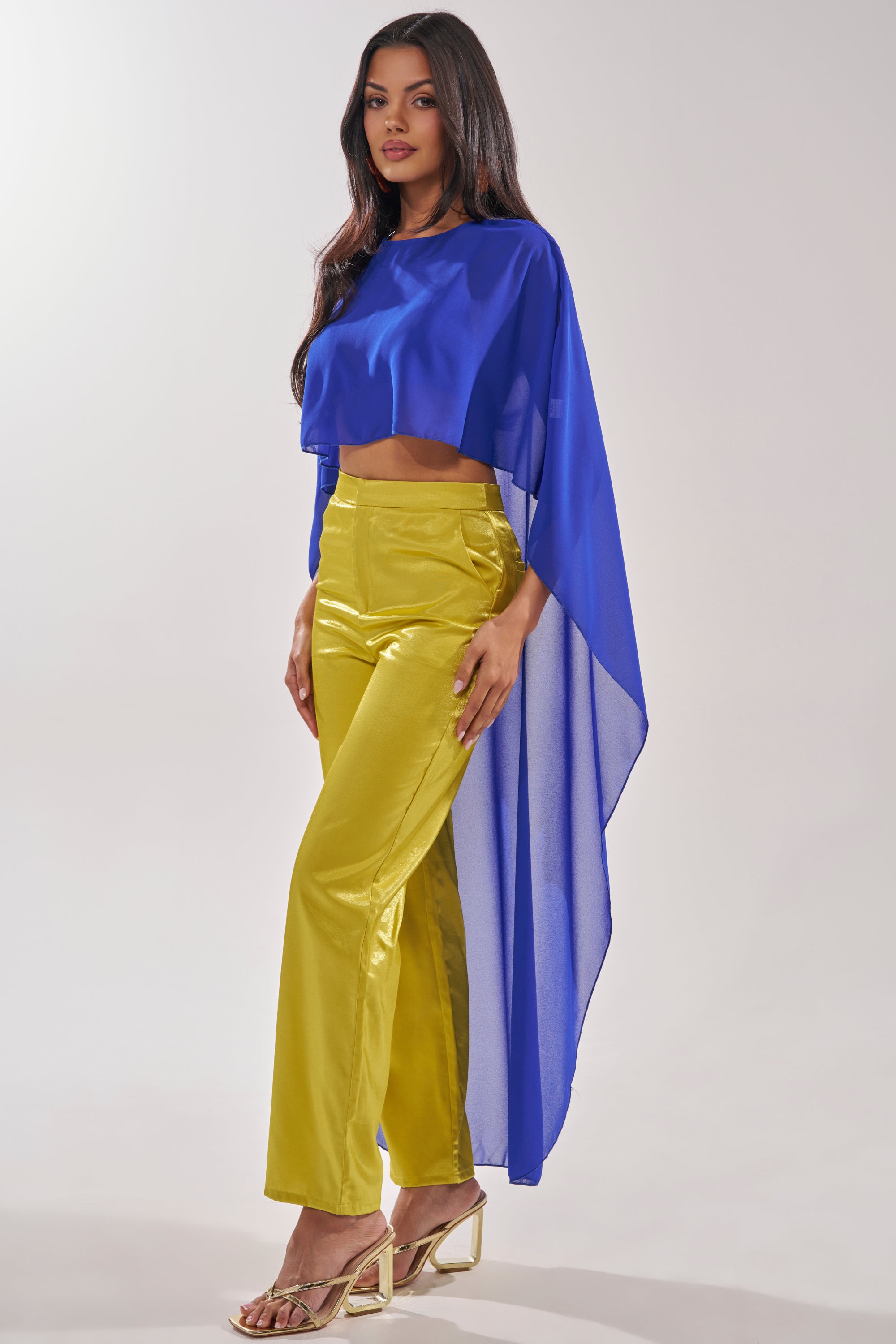 Against a neutral background, a woman models yellow satin pants, gold high-heeled sandals, and the COOL GIRL CHIFFON BLOUSE IN ROYAL BLUE featuring a sheer finish and an asymmetrical cape design. 5