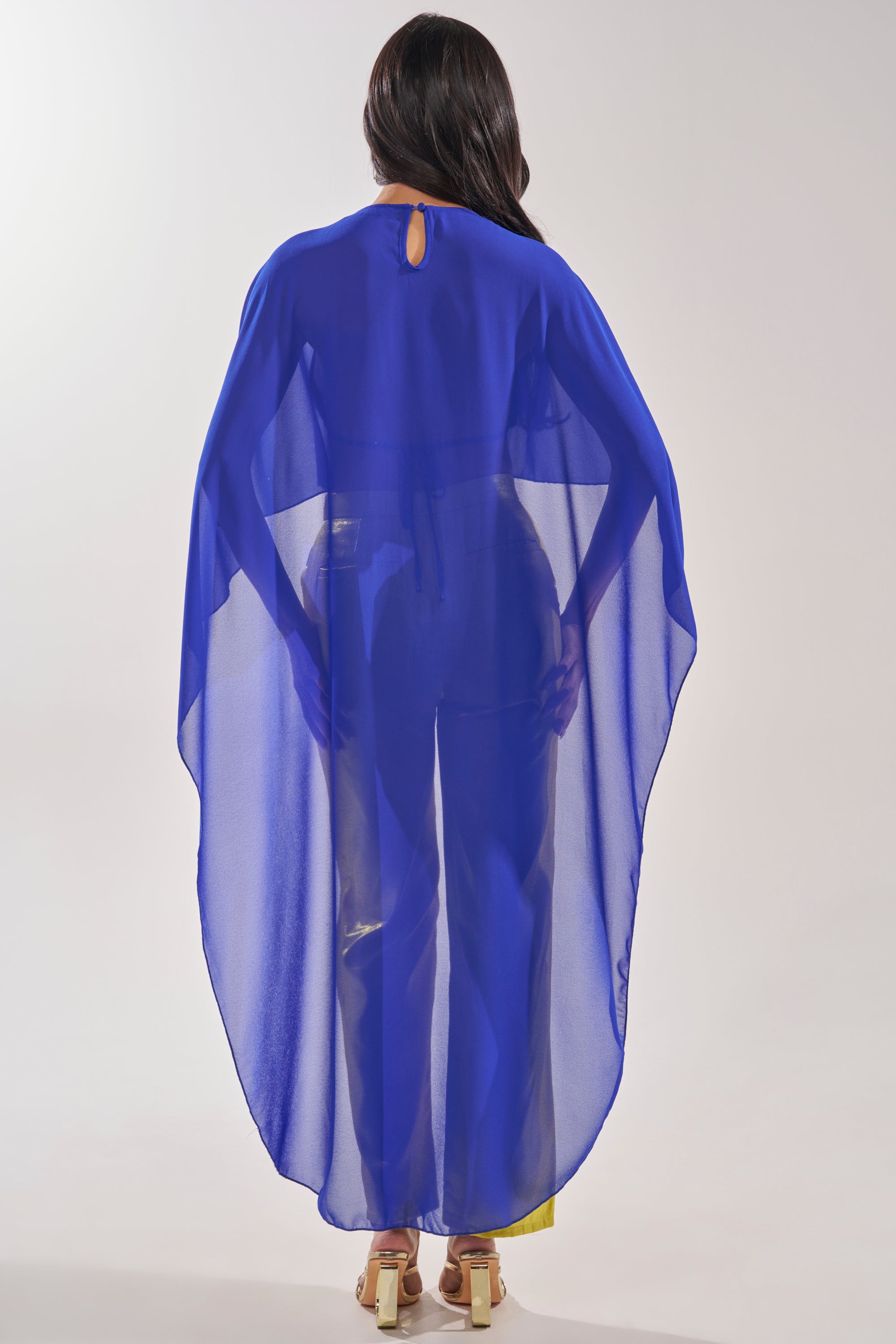 A woman faces away, wearing the COOL GIRL CHIFFON BLOUSE IN ROYAL BLUE as a cape-style top over matching pants and beige heels, set against a plain light background. 6