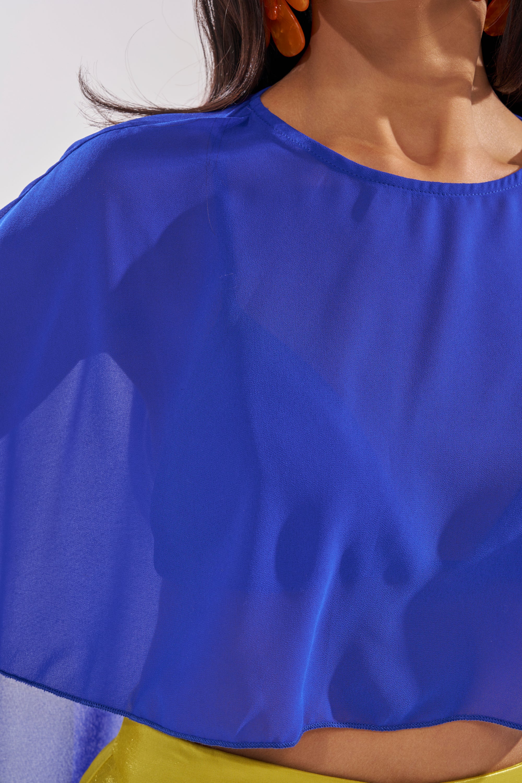 A person models the COOL GIRL CHIFFON BLOUSE IN ROYAL BLUE over a yellow top. The image is cropped to show only the lower face, neck, and torso, with orange earrings partly visible. 7