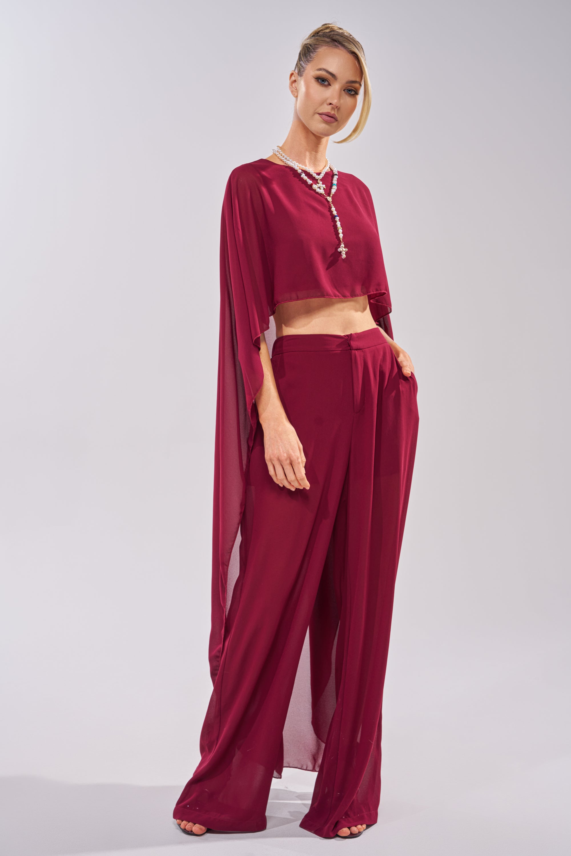 A woman wears the COOL GIRL CHIFFON BLOUSE IN BURGUNDY with matching wide-leg pants and a long necklace. Her hair is styled up, and she stands against a plain light gray background. 1